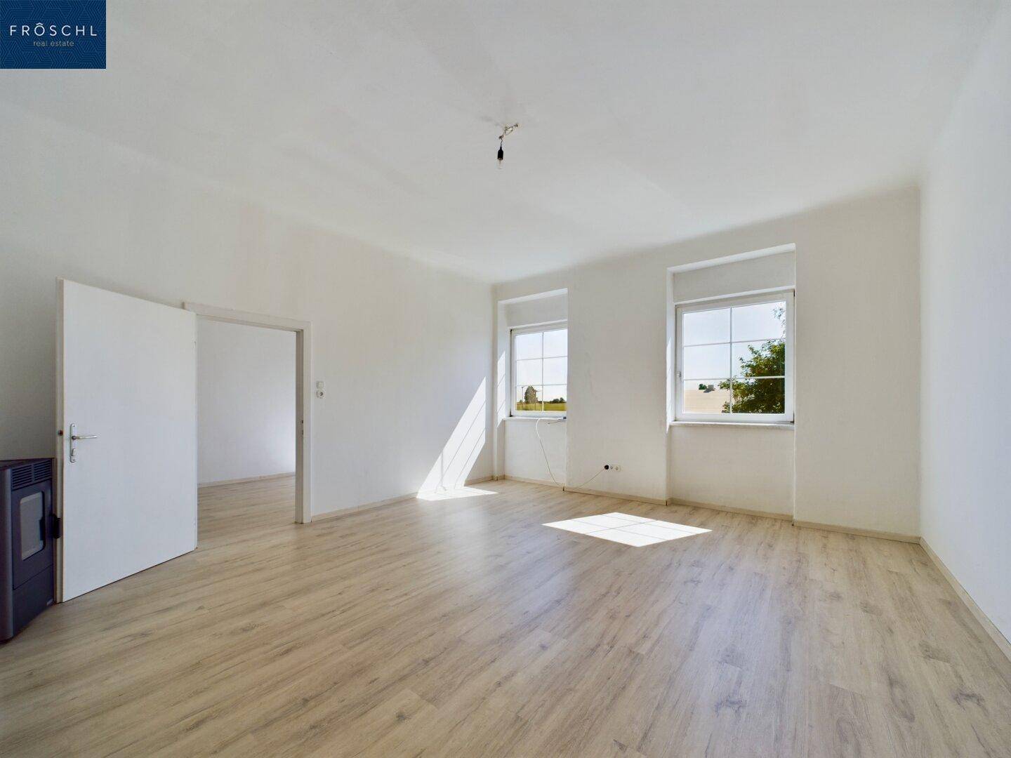 Geräumiges Wohnzimmer mit hellem Laminatboden und viel Tageslicht durch zwei große Fenster.