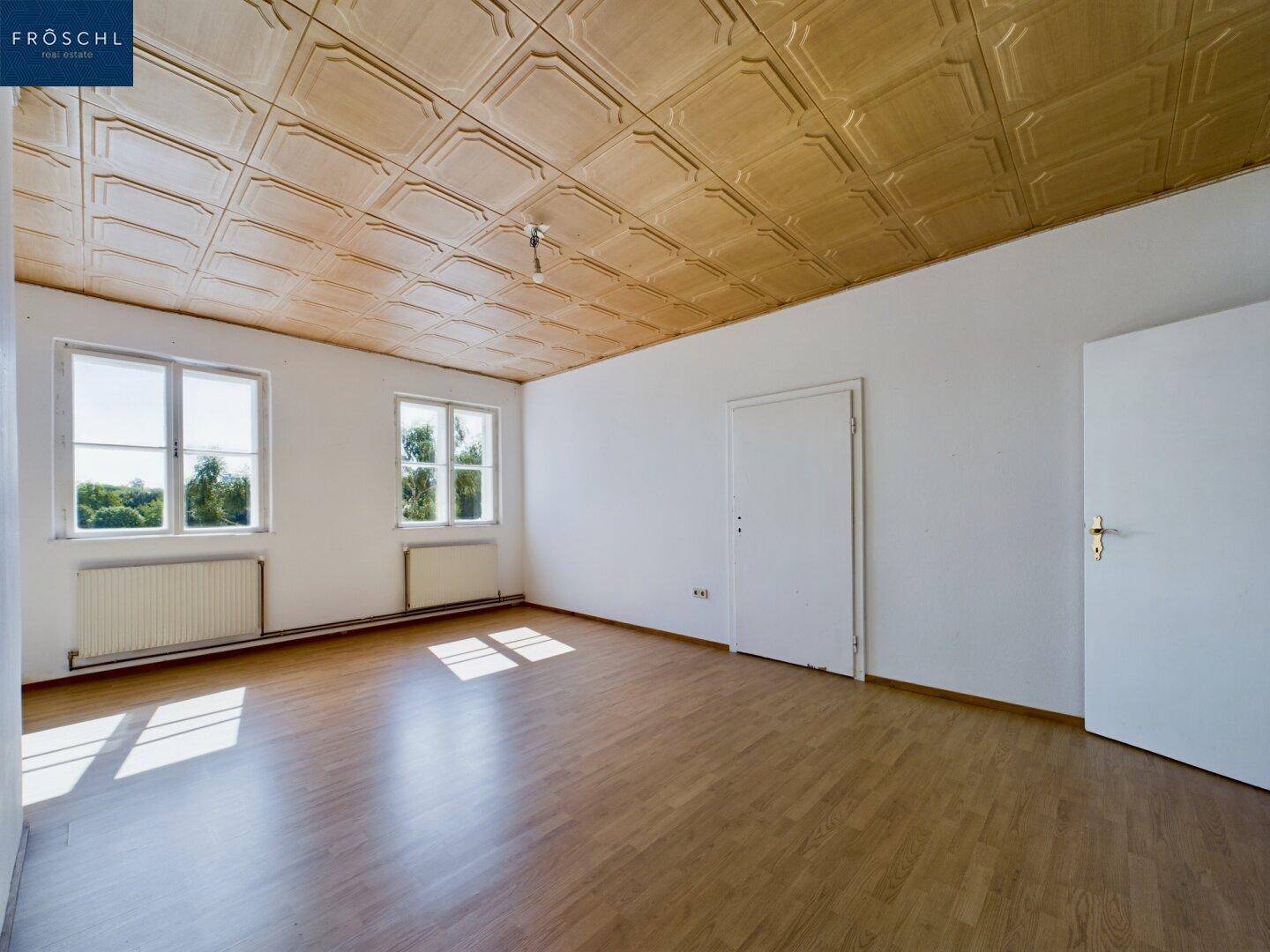 Heller Raum mit Holzboden, zwei Fenstern und einer dekorativen Deckenverkleidung.
