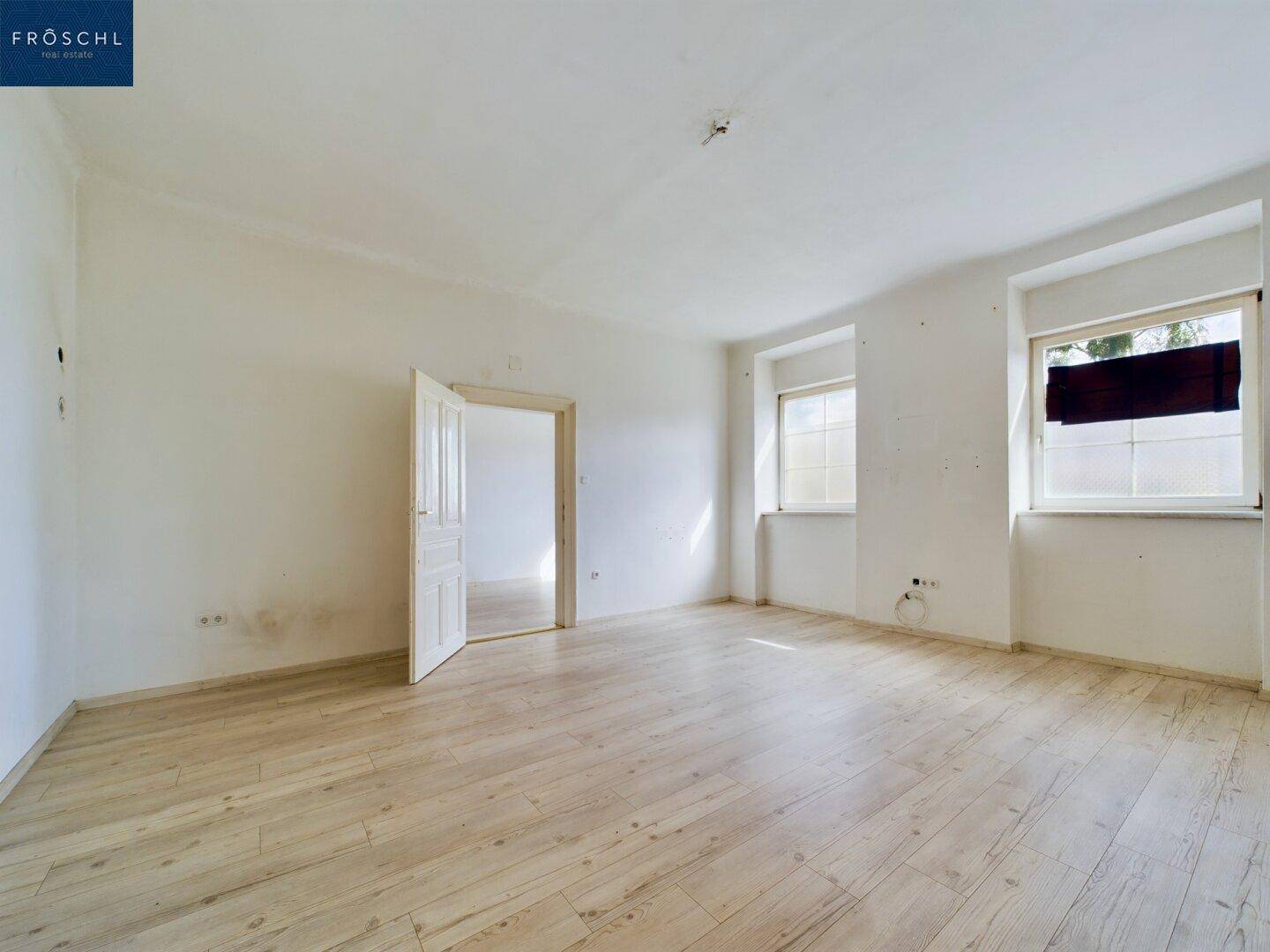 Geräumiger, heller Wohnraum mit Holzboden, zwei Fenstern und einer geöffneten Tür zu einem weiteren Zimmer.