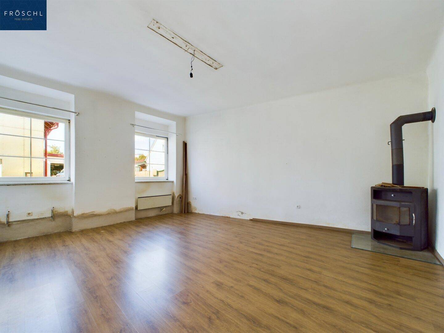 Heller Raum mit Holzboden, zwei Fenstern und einem modernen Holzofen als Wärmequelle.