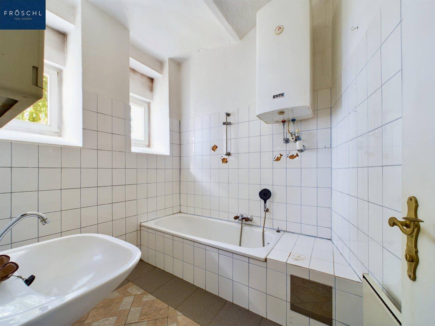 Badezimmer mit Badewanne, Waschbecken, weißen Fliesen und einem Warmwasserboiler an der Wand.