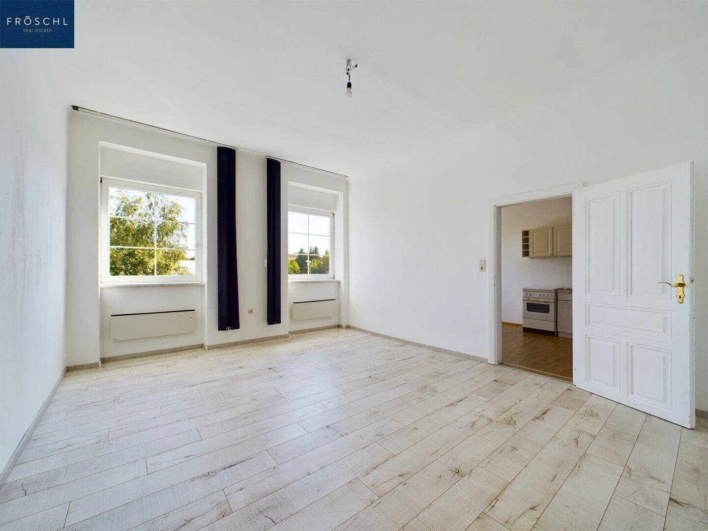 Heller Wohnraum mit Holzboden, zwei Fenstern und einer offenen Tür zu einem weiteren Zimmer.