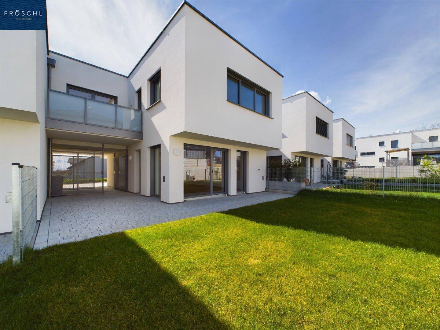 Rückseite der modernen Reihenhäuser mit gepflasterter Terrasse und grünem Rasen unter strahlendem Himmel.