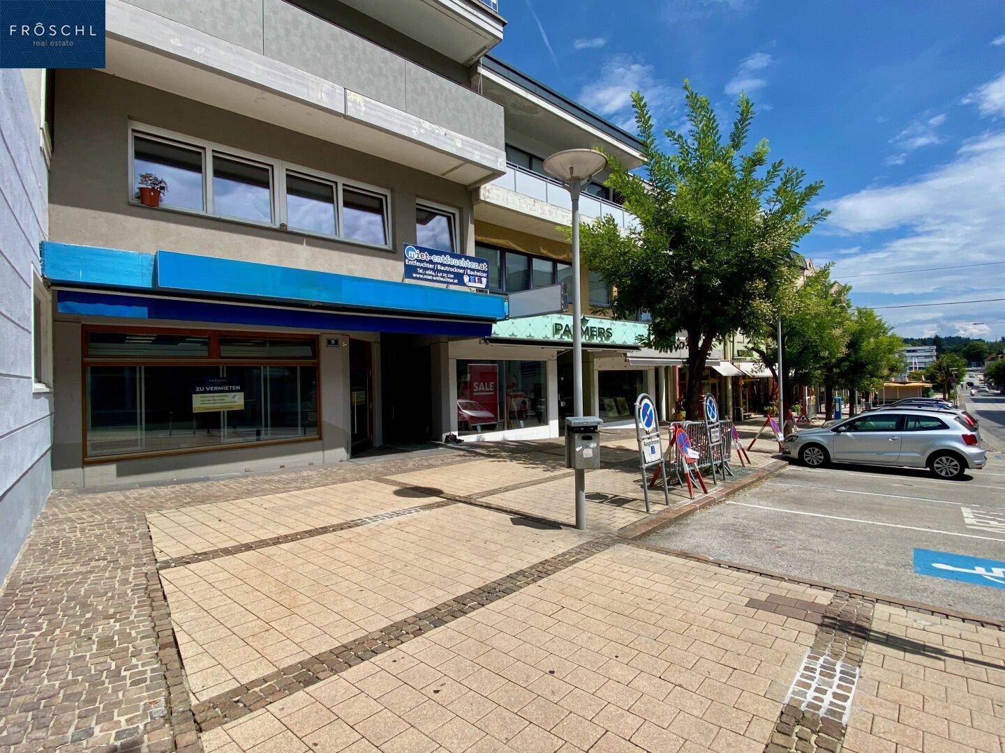 Fassade eines Geschäftsgebäudes mit Schaufenstern, Bürgersteig und geparkten Autos auf der Straße.