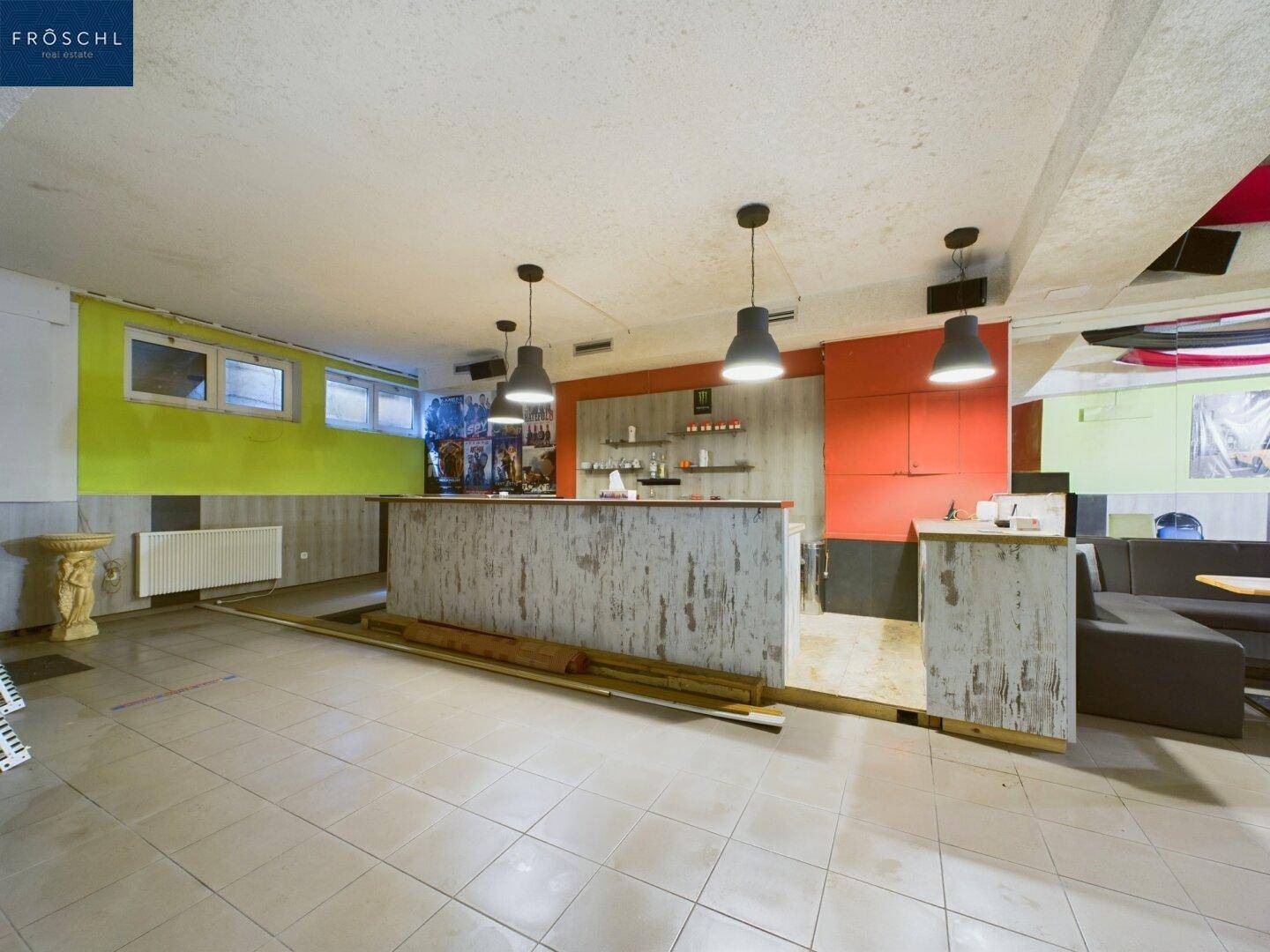 Bar-Bereich mit rustikaler Holztheke, Fliesenboden und farbigen Wänden in einem Gastraum.