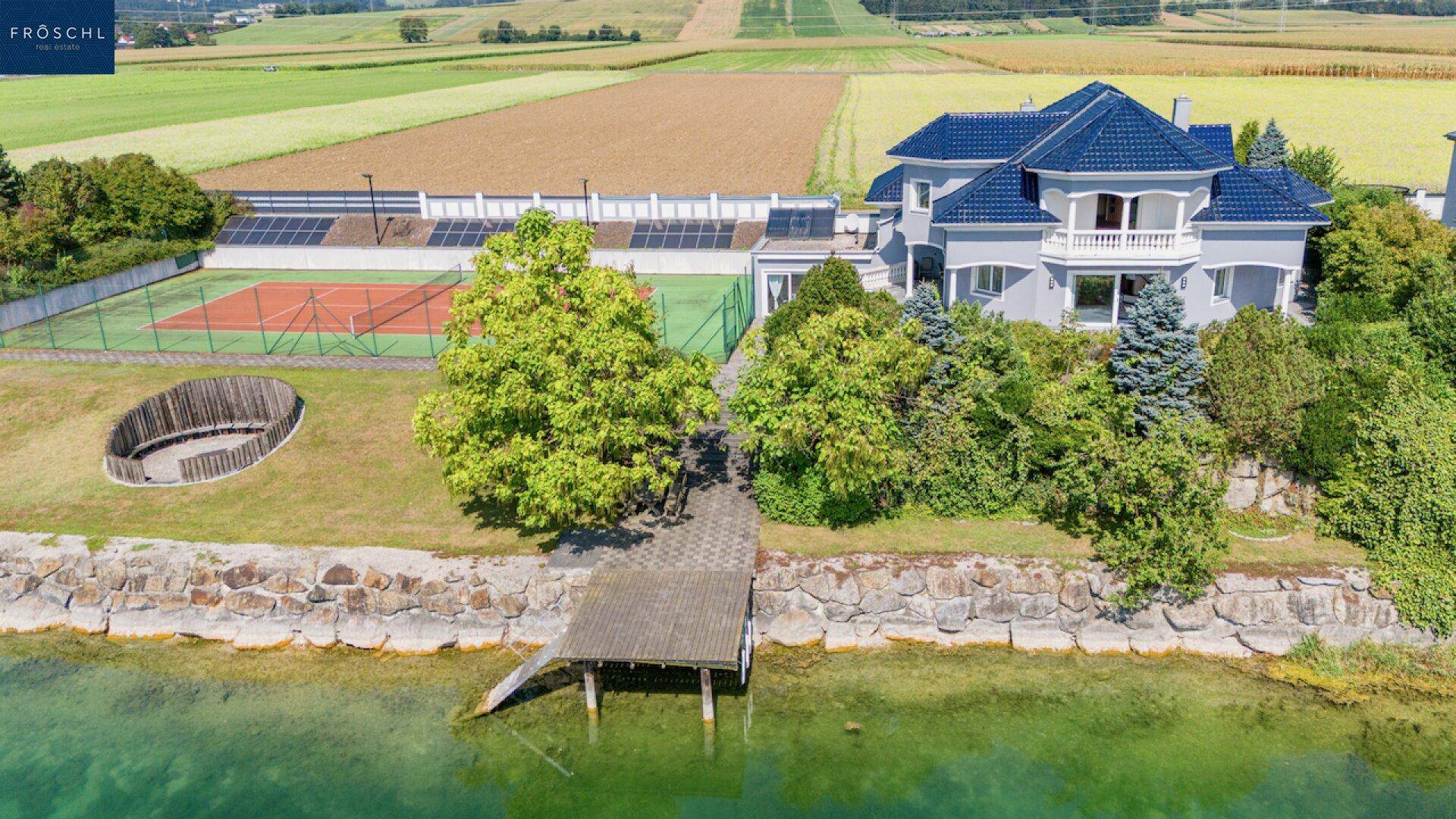 Weitläufige Ansicht des Anwesens mit Seezugang, Tennisplatz und umliegenden Feldern.