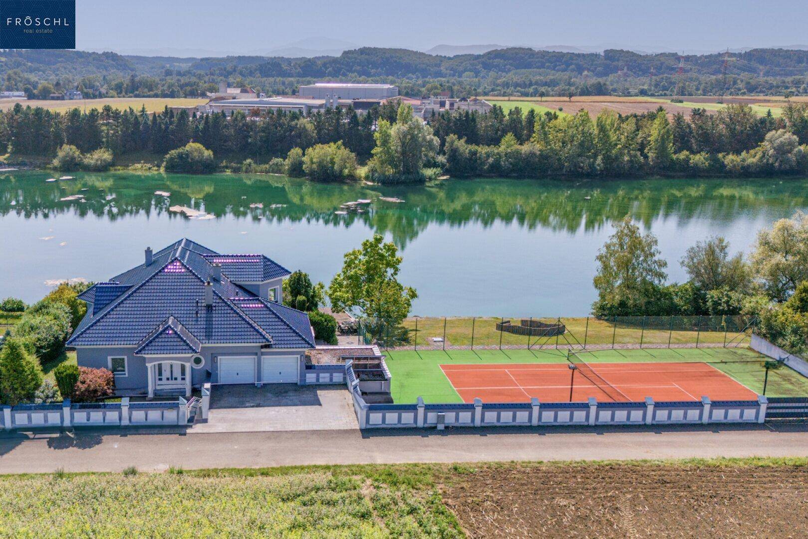 Luftaufnahme eines Hauses am See mit eigenem Tennisplatz und Doppelgarage.