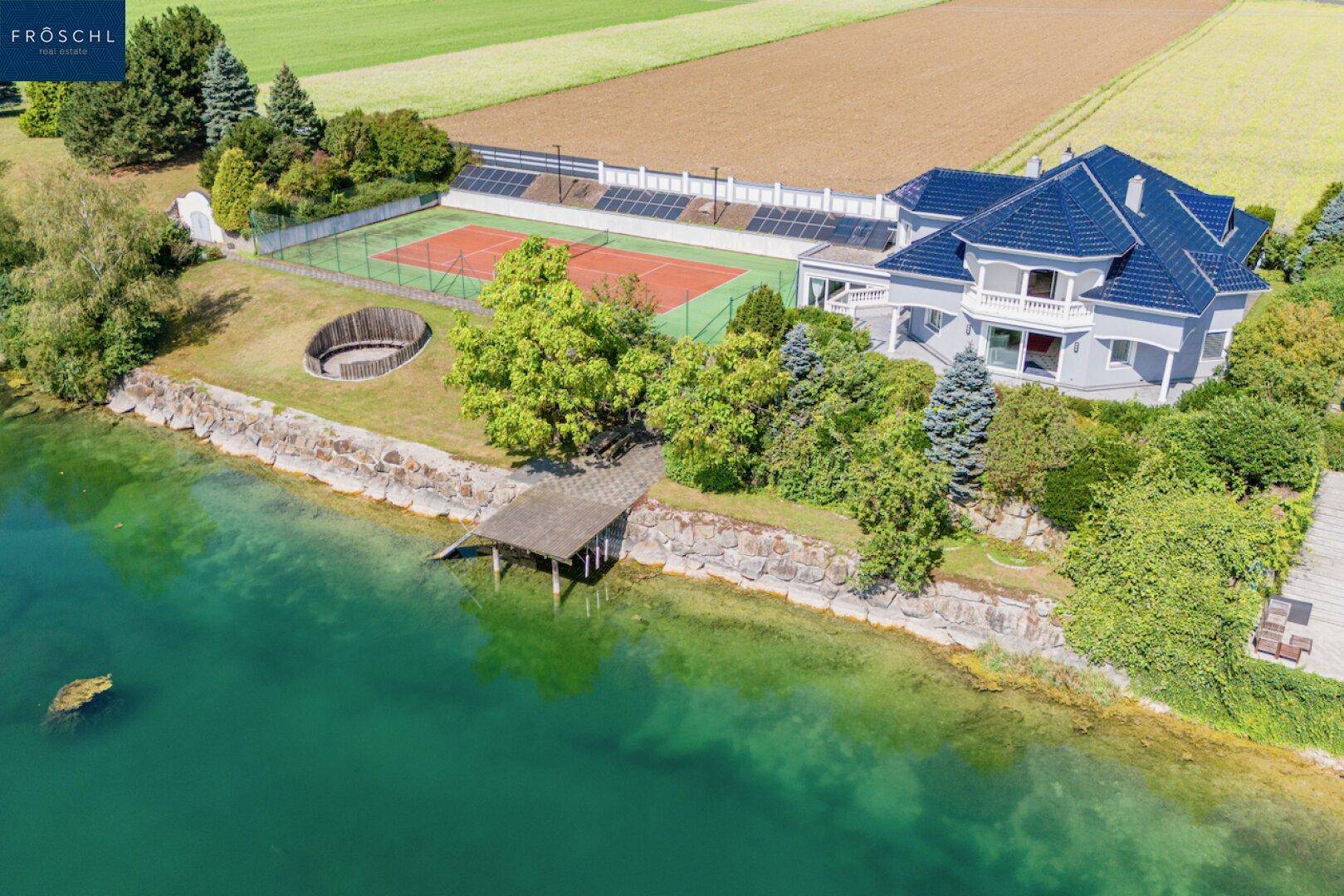 Weitläufige Ansicht des Anwesens mit Seezugang, Tennisplatz und umliegenden Feldern.