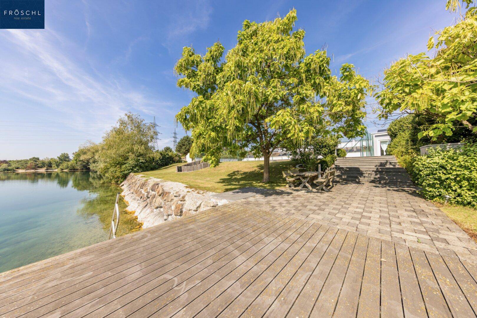 Holzterrasse am Seeufer mit Picknicktisch und altem Baum, ideal für Entspannung.