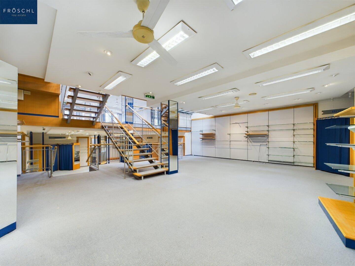 Weitläufiger Verkaufsraum mit Teppichboden, Deckenventilatoren und einem Blick auf die Treppen und Ausstellungsflächen.