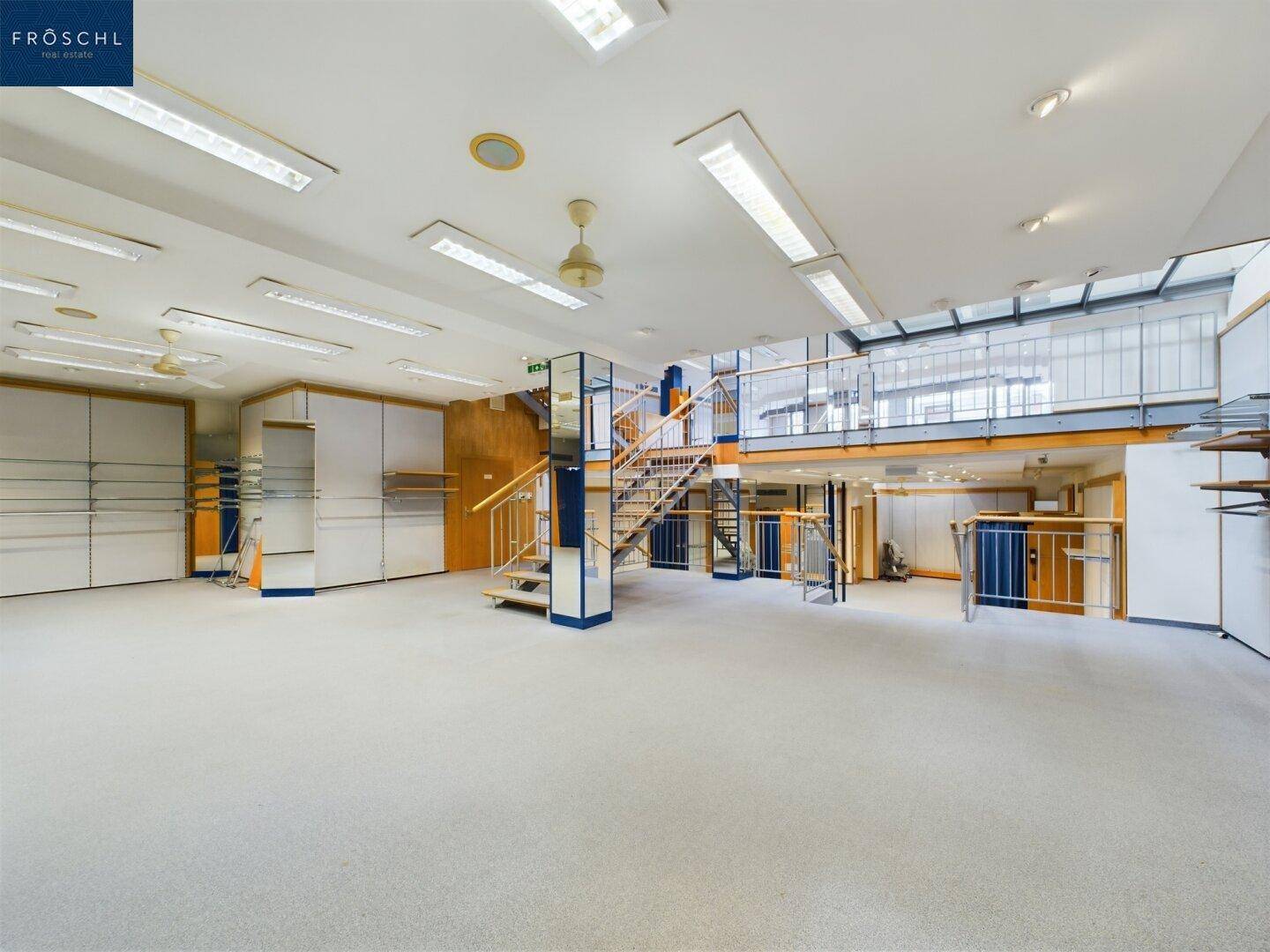 Großer, offener Verkaufsraum mit Teppichboden, Deckenbeleuchtung und einer Galerie, die verschiedene Bereiche verbindet.