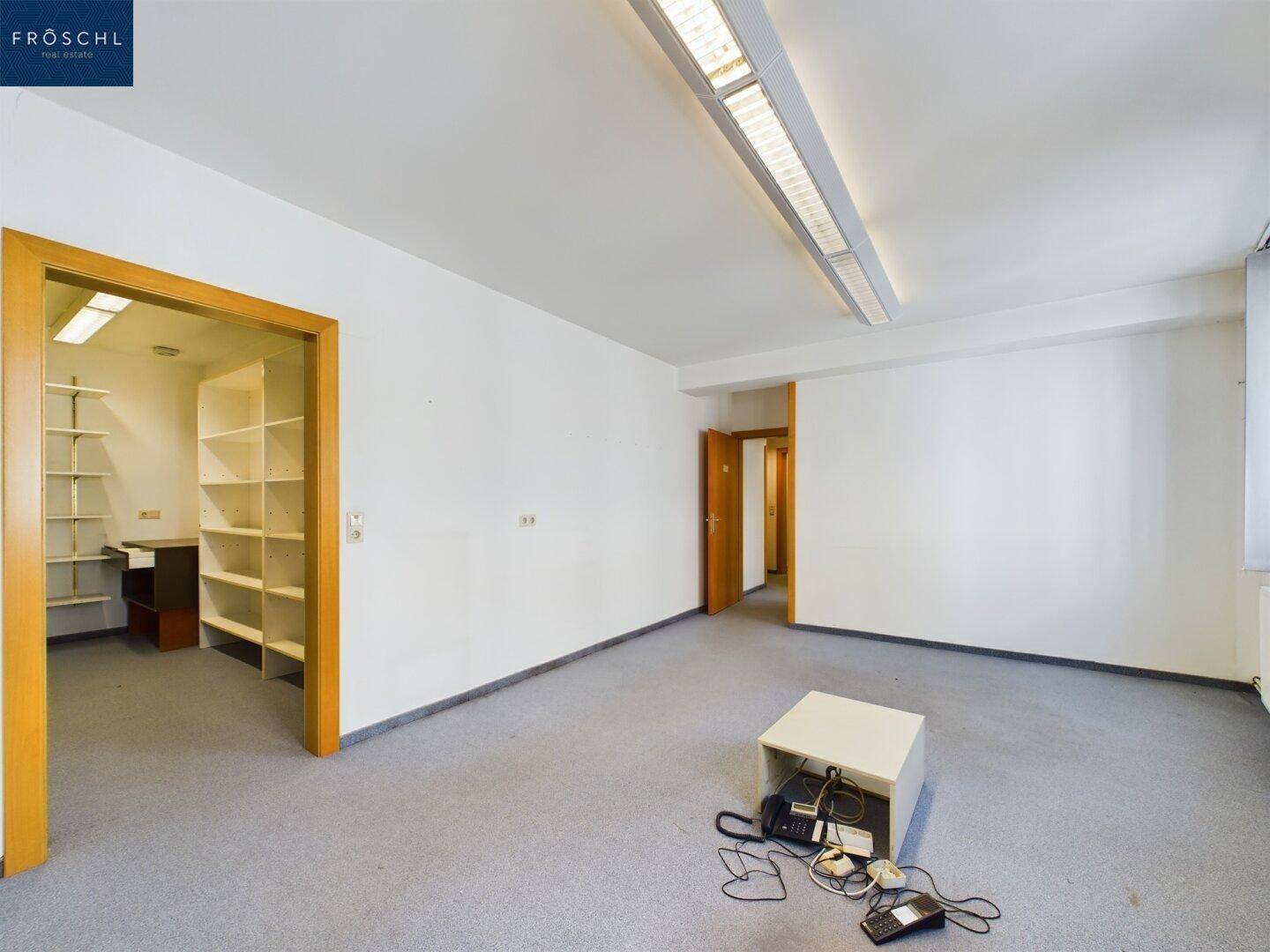 Leeres Büro mit Teppichboden und einem offenen Durchgang zu einem Raum mit Regalen.