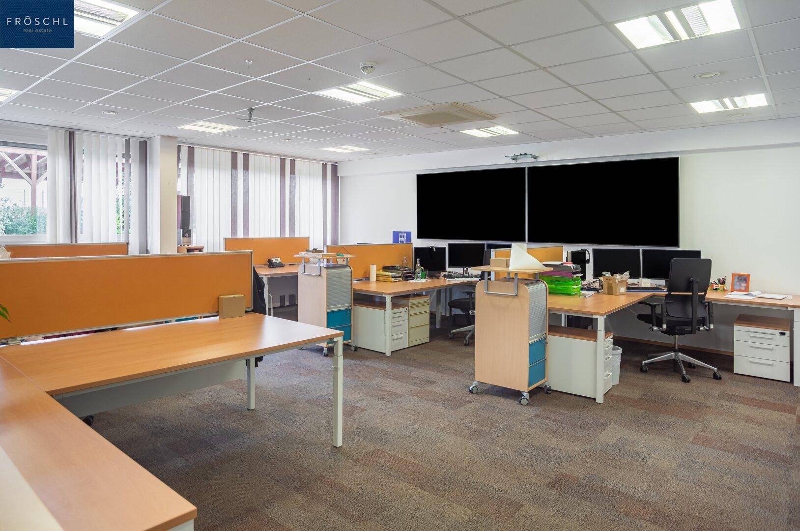 Modernes Großraumbüro mit vielen Arbeitsplätzen, Computern und einem großen Bildschirm an der Wand.