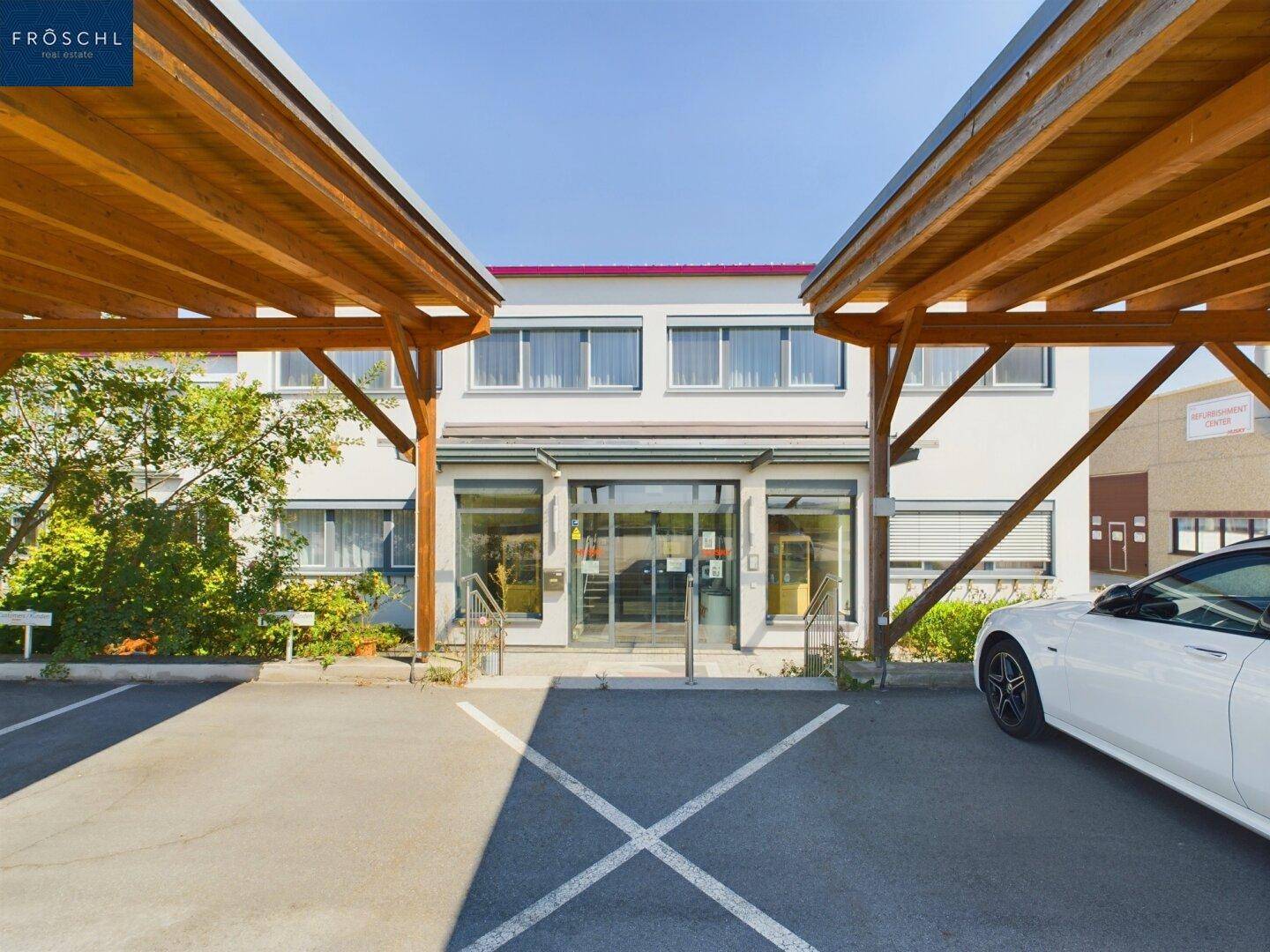 Eingangsbereich eines modernen Bürogebäudes mit überdachten Parkplätzen im Vordergrund.