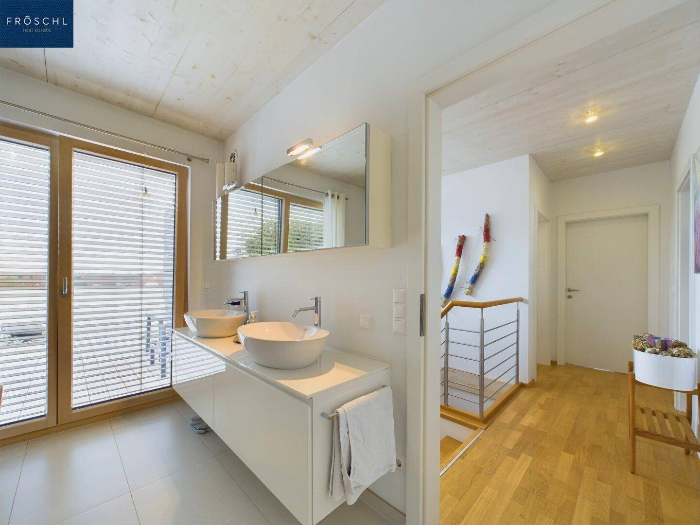 Modernes Badezimmer mit Doppelwaschbecken und großem Fenster, angrenzend an einen hellen Flur.