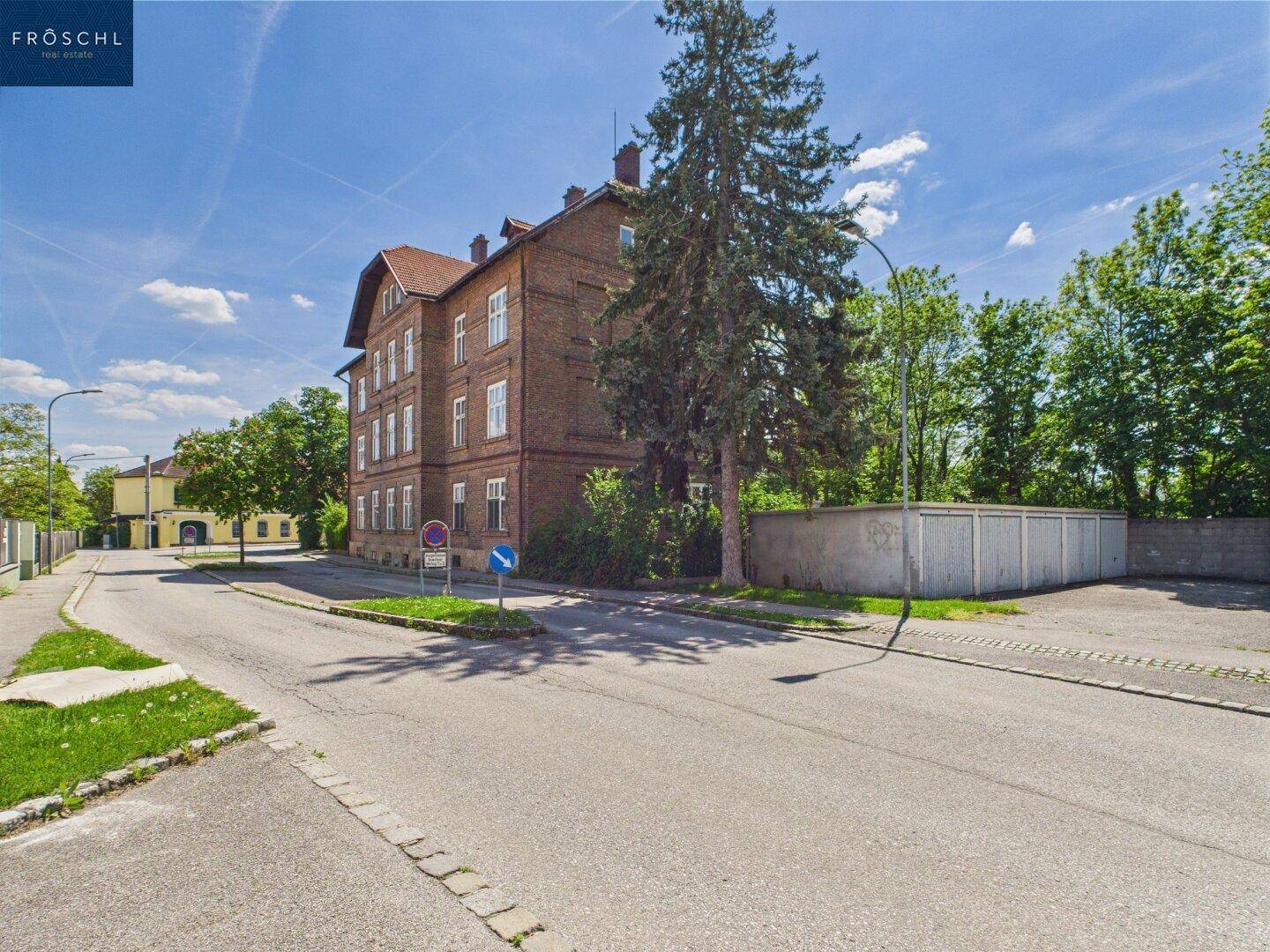 Seitenansicht eines Ziegelgebäudes an einer Straße mit Bäumen und einer Garagenreihe im Hintergrund.