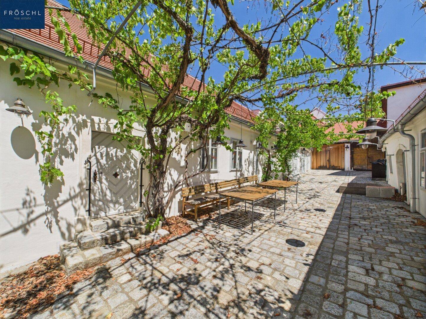Sonniger Innenhof mit Kopfsteinpflaster und rustikalen Holzbänken unter Weinreben.