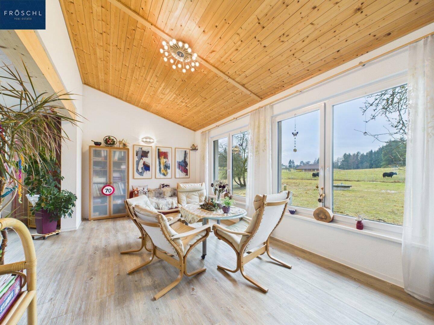 Heller Wintergarten mit Holzdecke, großen Fenstern und gemütlicher Sitzecke mit Blick ins Grüne.