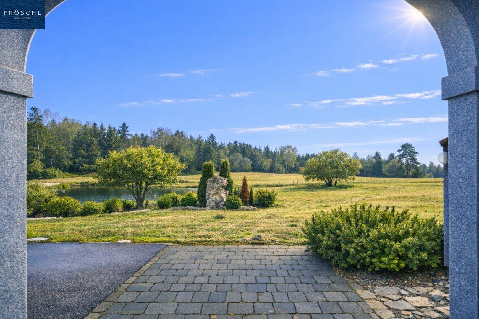 Gepflasterter Eingangsbereich mit weitem Blick über grüne Wiesen, Bäume und einen kleinen Teich.