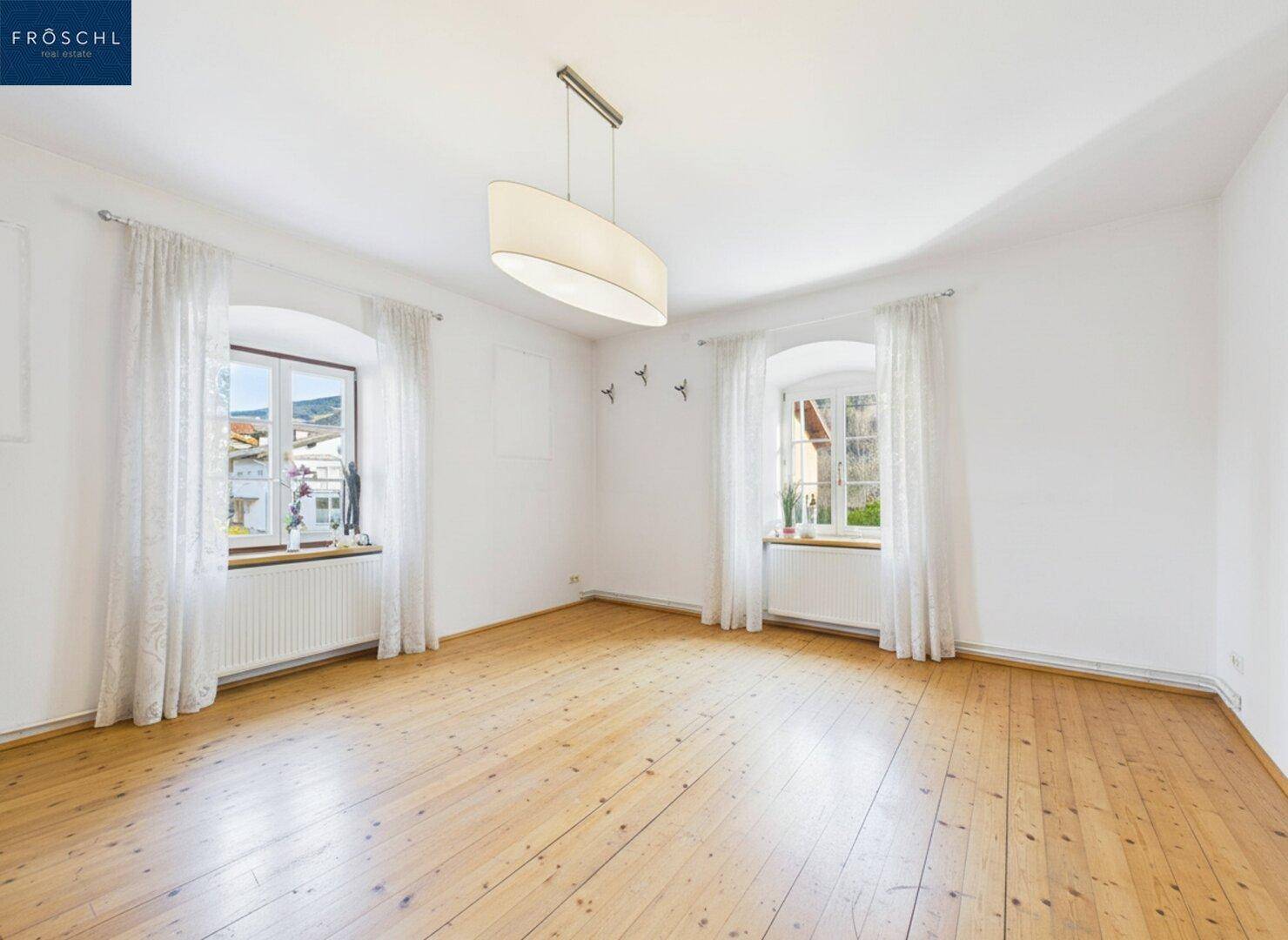Geräumiges Wohnzimmer mit Holzdielenboden, hohen Decken und zwei Fenstern.
