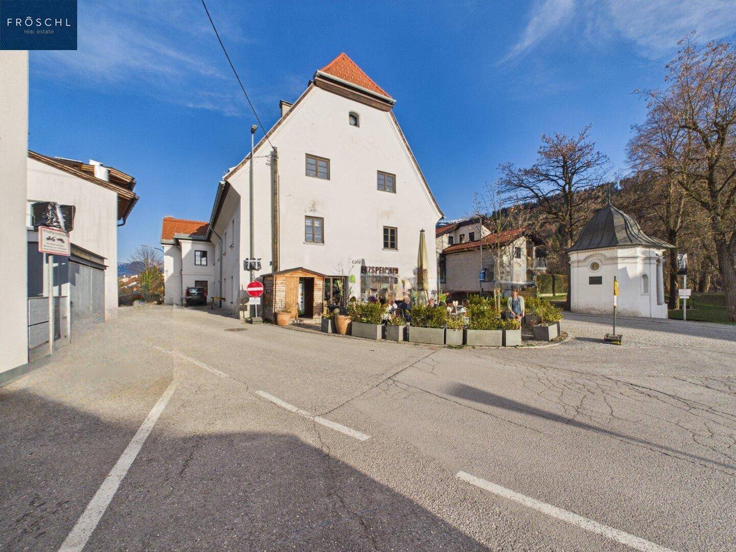 Historisches Gebäude an einer Kreuzung mit Außengastronomie und Kapelle im Hintergrund.