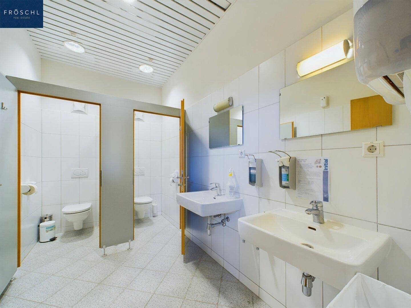 Öffentlicher Waschraum mit mehreren Toilettenkabinen, Waschbecken und hell gefliesten Wänden und Böden.