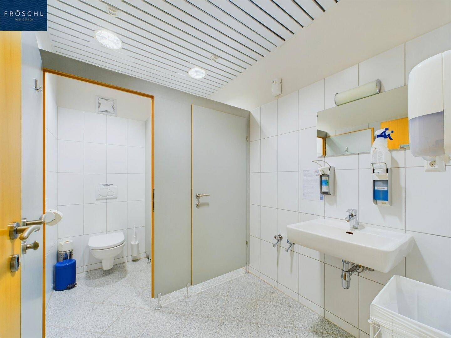 Heller und sauberer Sanitärbereich mit weißen Fliesen, mehreren Toilettenkabinen und Waschbecken.