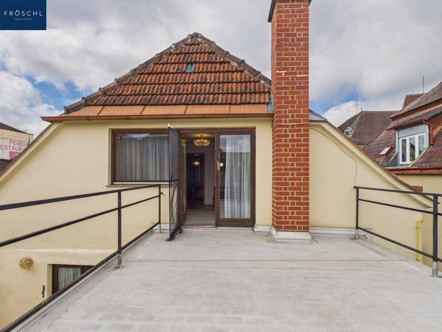 Dachterrasse mit Ziegelkamin und Zugang zu einem Innenraum, umgeben von Dächern.