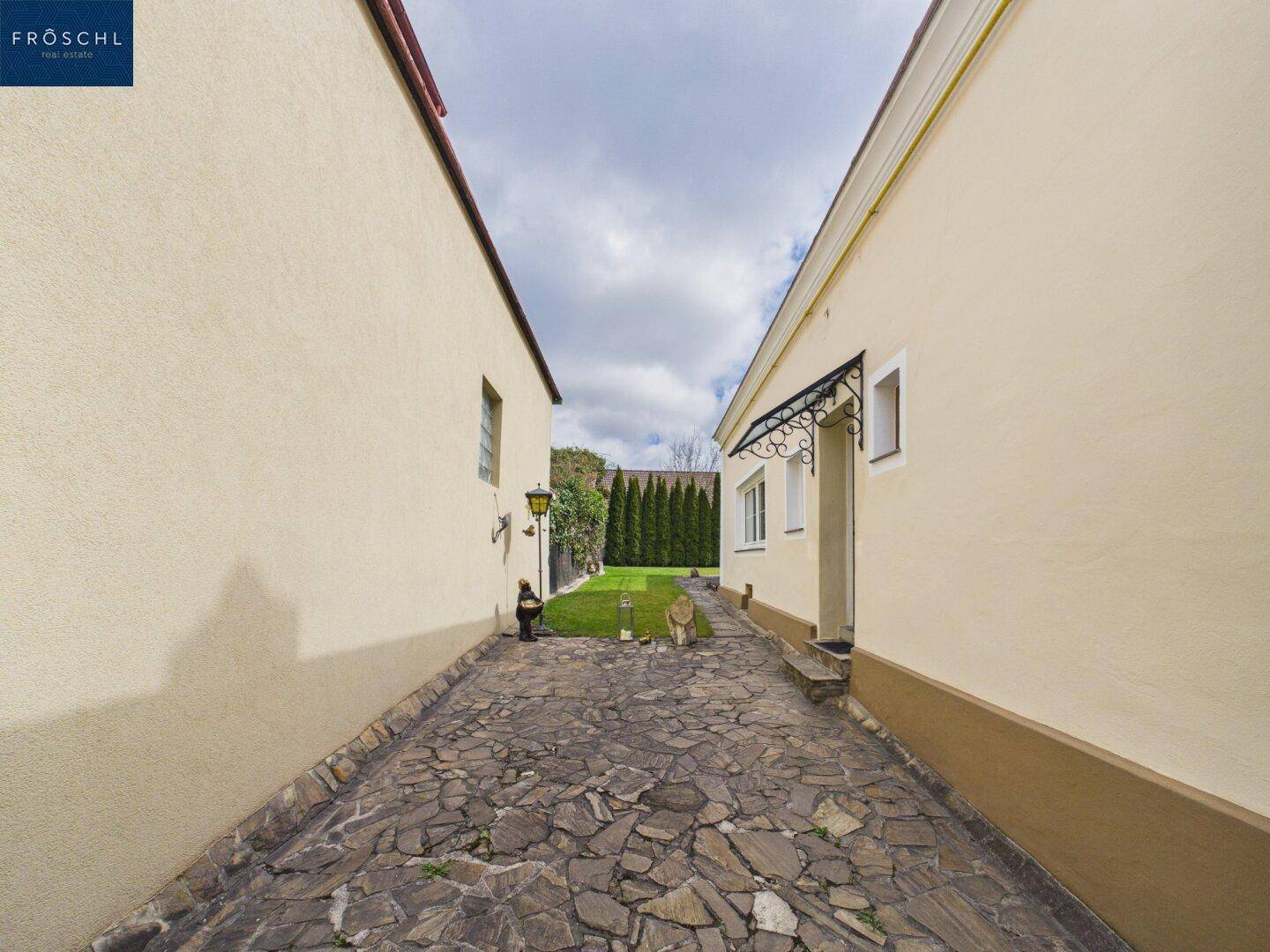 Steiniger Zufahrtsweg zwischen zwei Gebäuden, führt zu einem grünen Gartenbereich.