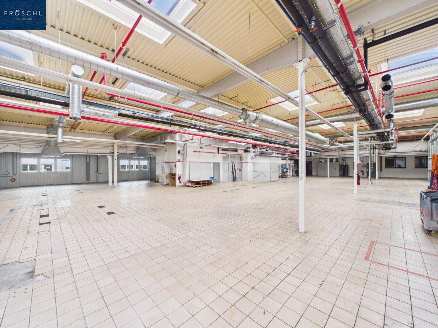 Weitläufige Industriehalle mit Fliesenboden, hohen Decken und zahlreichen sichtbaren Rohrleitungen und Lüftungsschächten.
