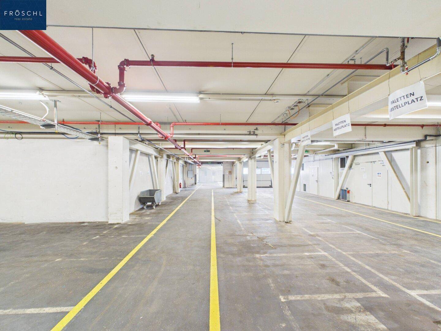 Industrieller Bereich mit gelben Bodenmarkierungen, Betonboden und sichtbaren roten Rohrleitungen an der Decke.