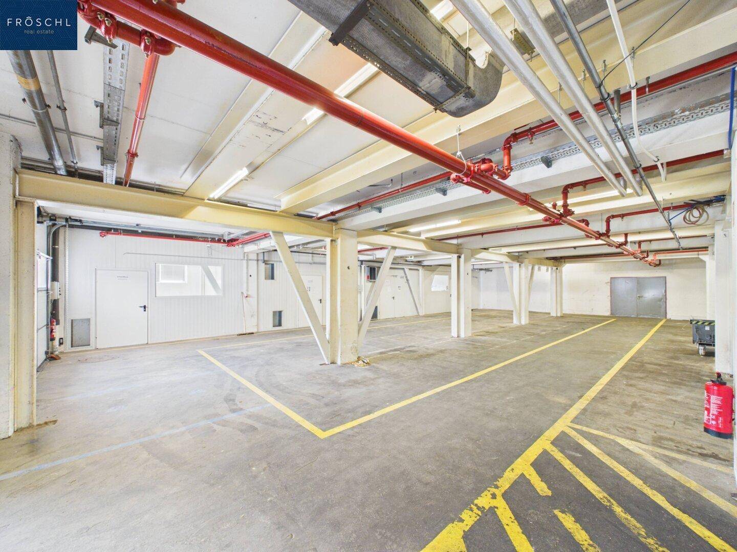 Großer Industrieraum mit Betonboden, gelben Markierungen und komplexen Rohrleitungen an der Decke.