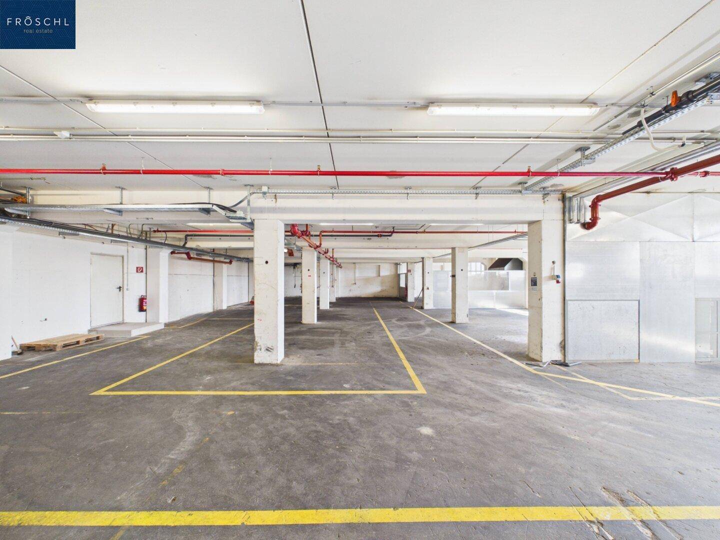 Tiefgarage mit Betonboden, gelben Parkplatzmarkierungen und tragenden Säulen sowie roten Rohrleitungen.