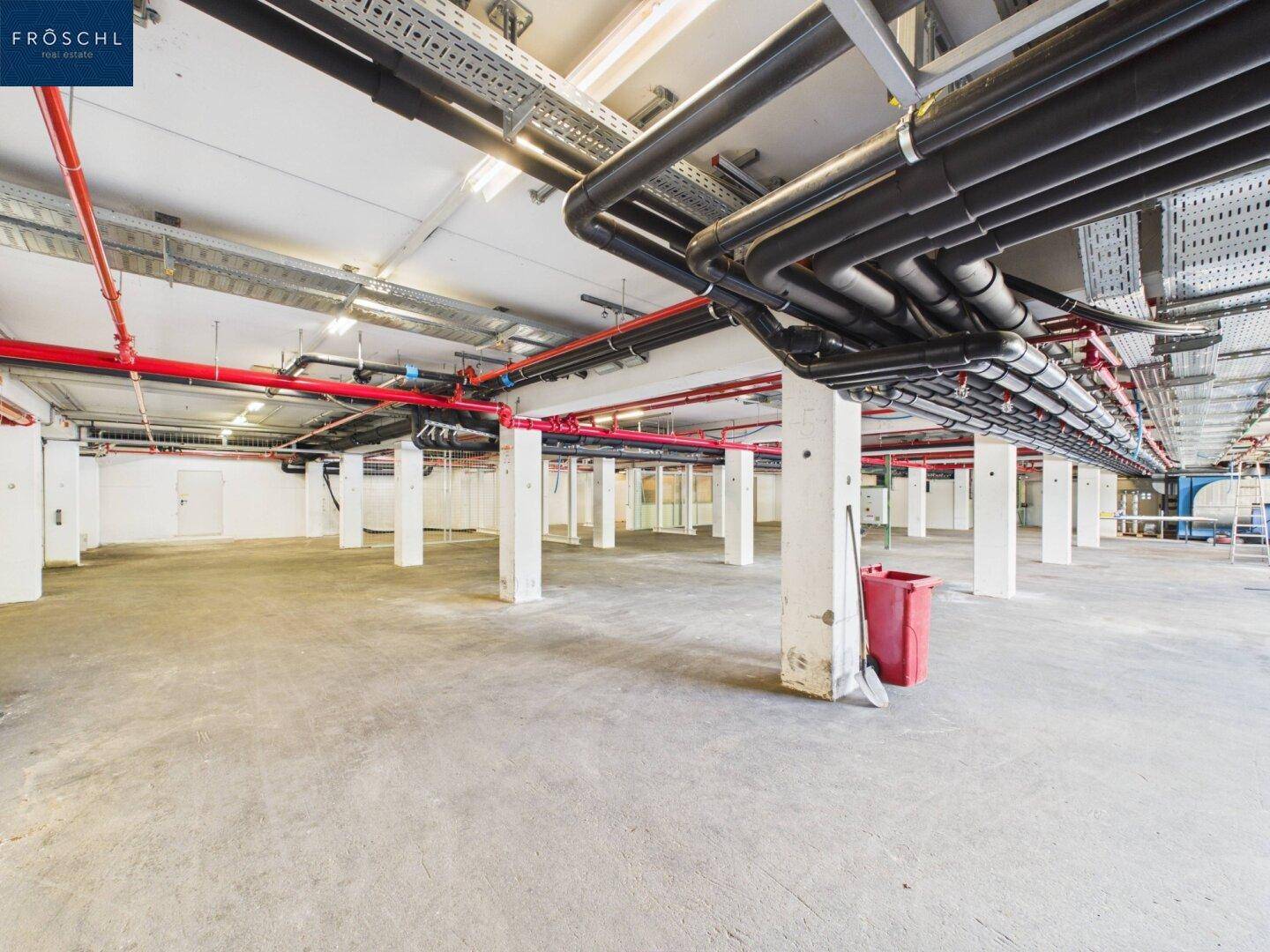 Industrieraum mit Betonboden, Säulen und einem dichten Netz aus schwarzen und roten Rohrleitungen an der Decke.