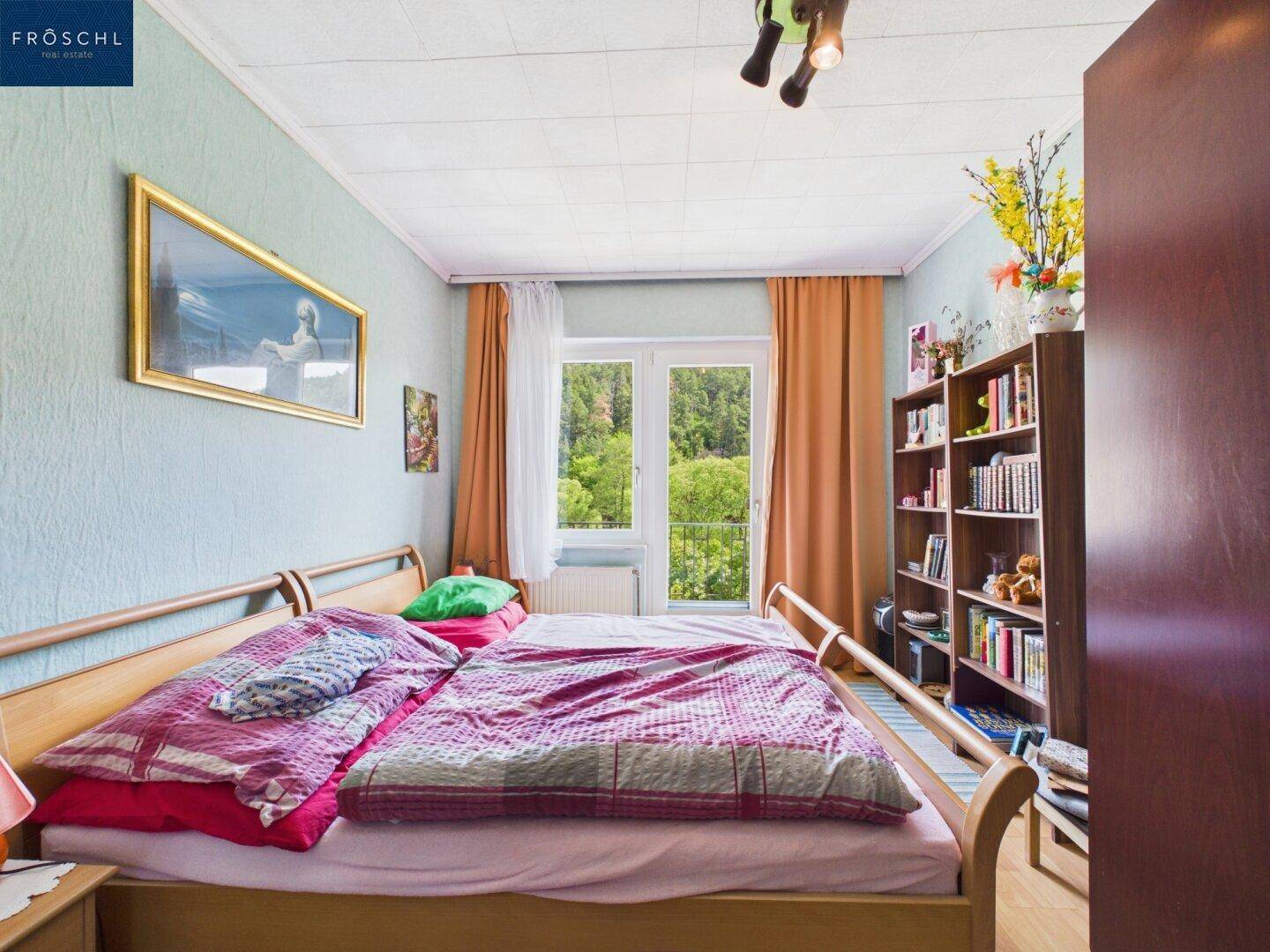 Schlafzimmer mit Doppelbett, Bücherregal und Blick ins Grüne durch große Fenster.
