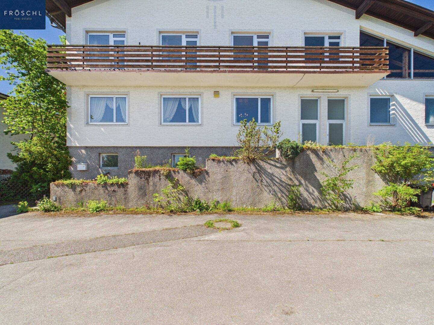 Vorderansicht eines Wohnhauses mit Balkon und gepflastertem Vorplatz an einem sonnigen Tag.