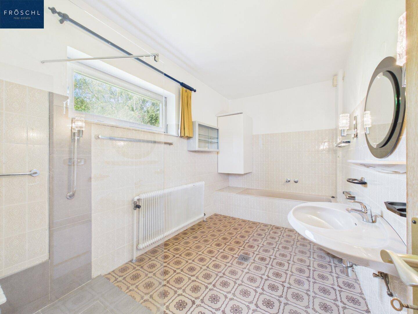 Badezimmer mit Badewanne, Dusche, Fenster und gemusterten Fliesen.