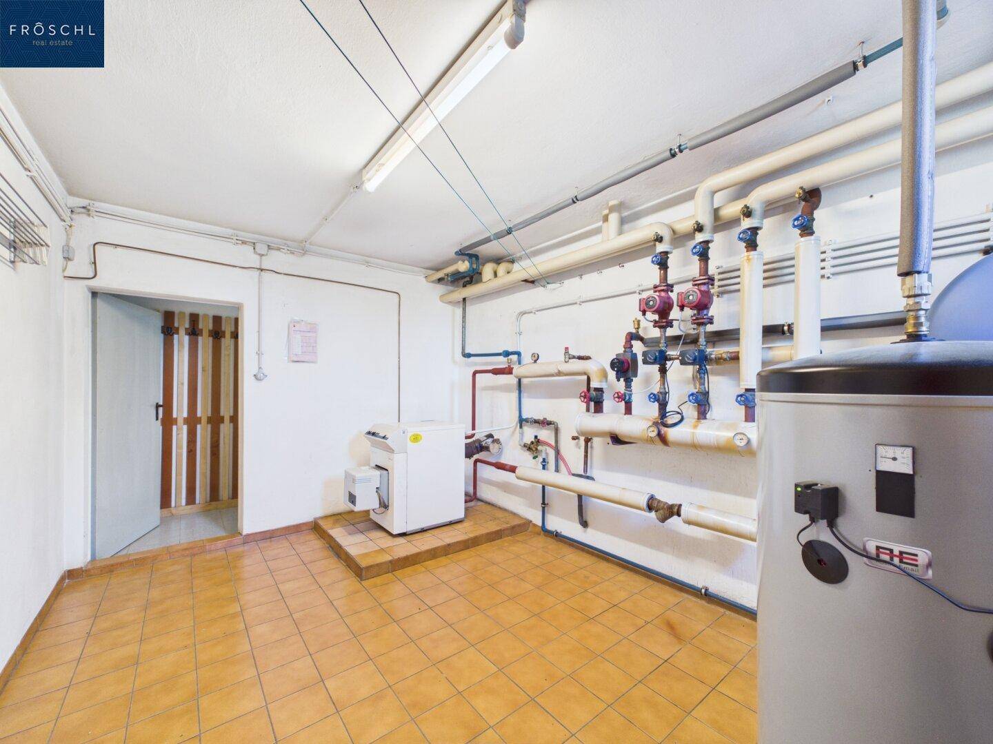 Heizungsraum im Keller mit Warmwasserspeicher und komplexer Rohrinstallation auf Fliesenboden.