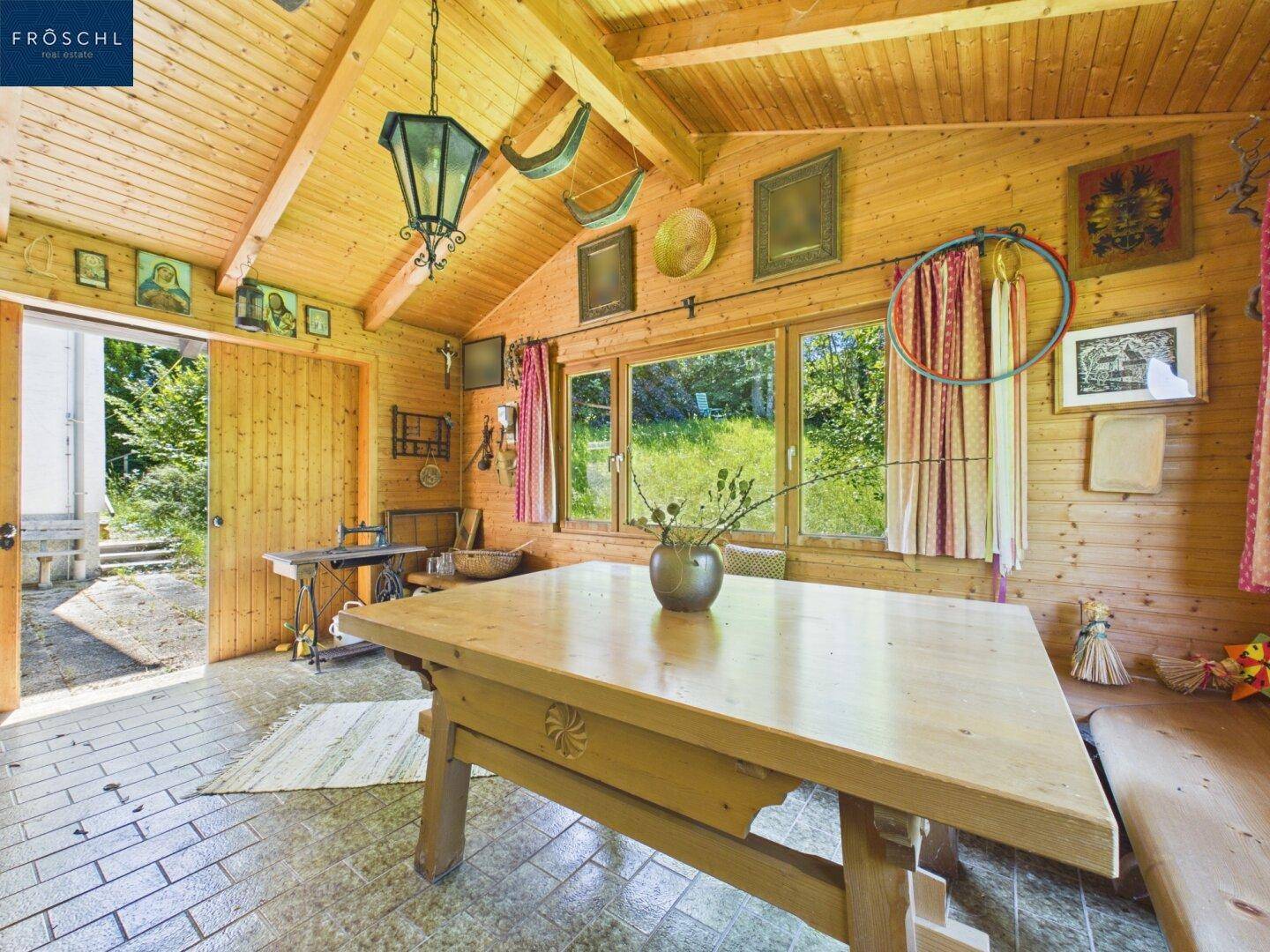 Gemütlicher Holzpavillon mit großen Fenstern, rustikalem Holztisch und Blick ins Grüne.