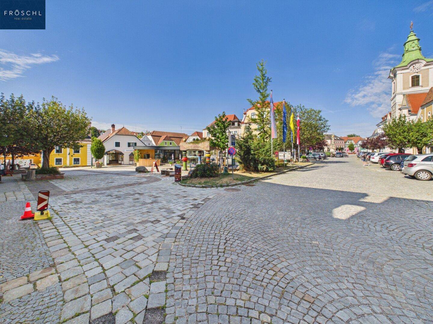 Weiter Blick über einen gepflasterten Stadtplatz mit Bäumen, Autos und einem Kirchturm im Hintergrund.
