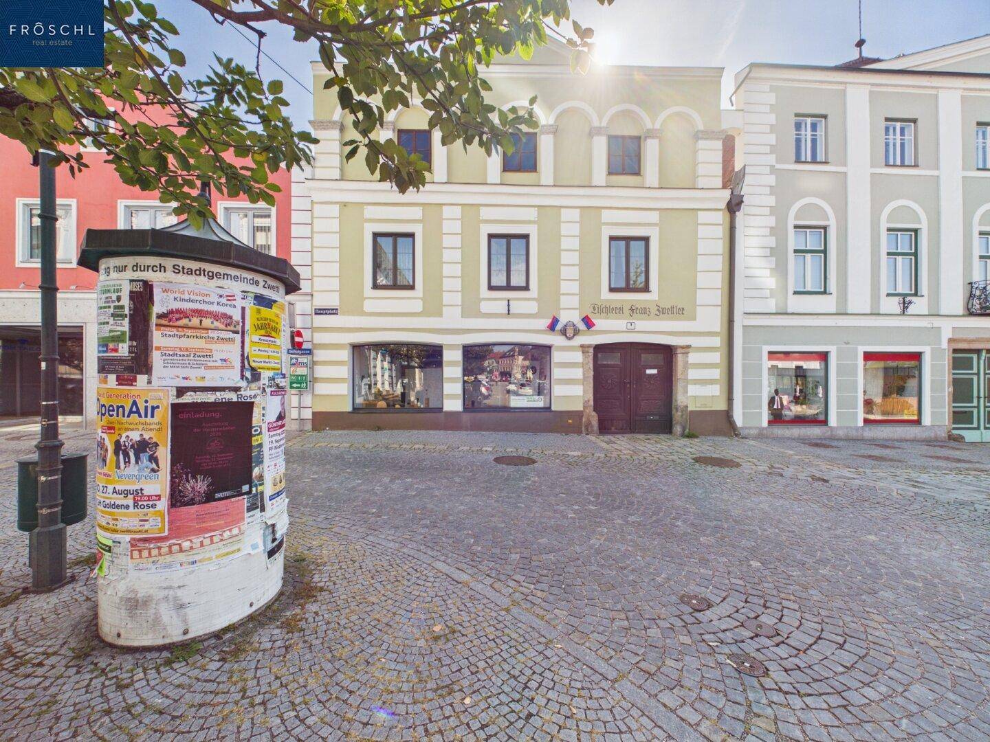Historisches Gebäude mit Geschäften im Erdgeschoss an einer gepflasterten Straße mit Litfaßsäule.