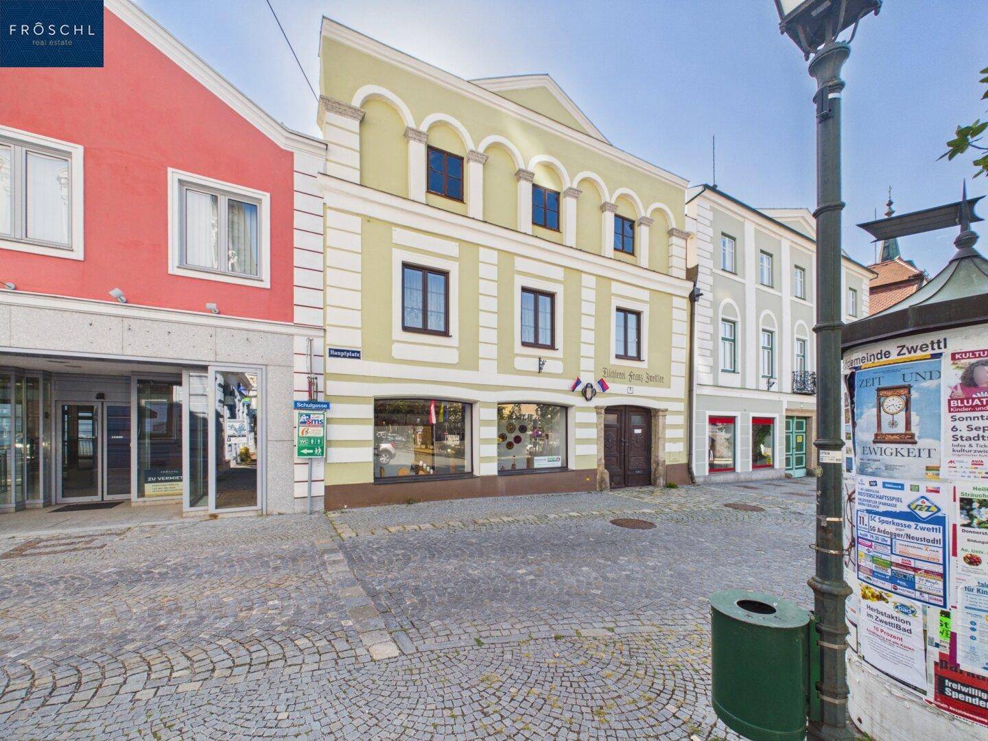 Historisches Gebäude mit gelber Fassade und Geschäften im Erdgeschoss an einem gepflasterten Platz.