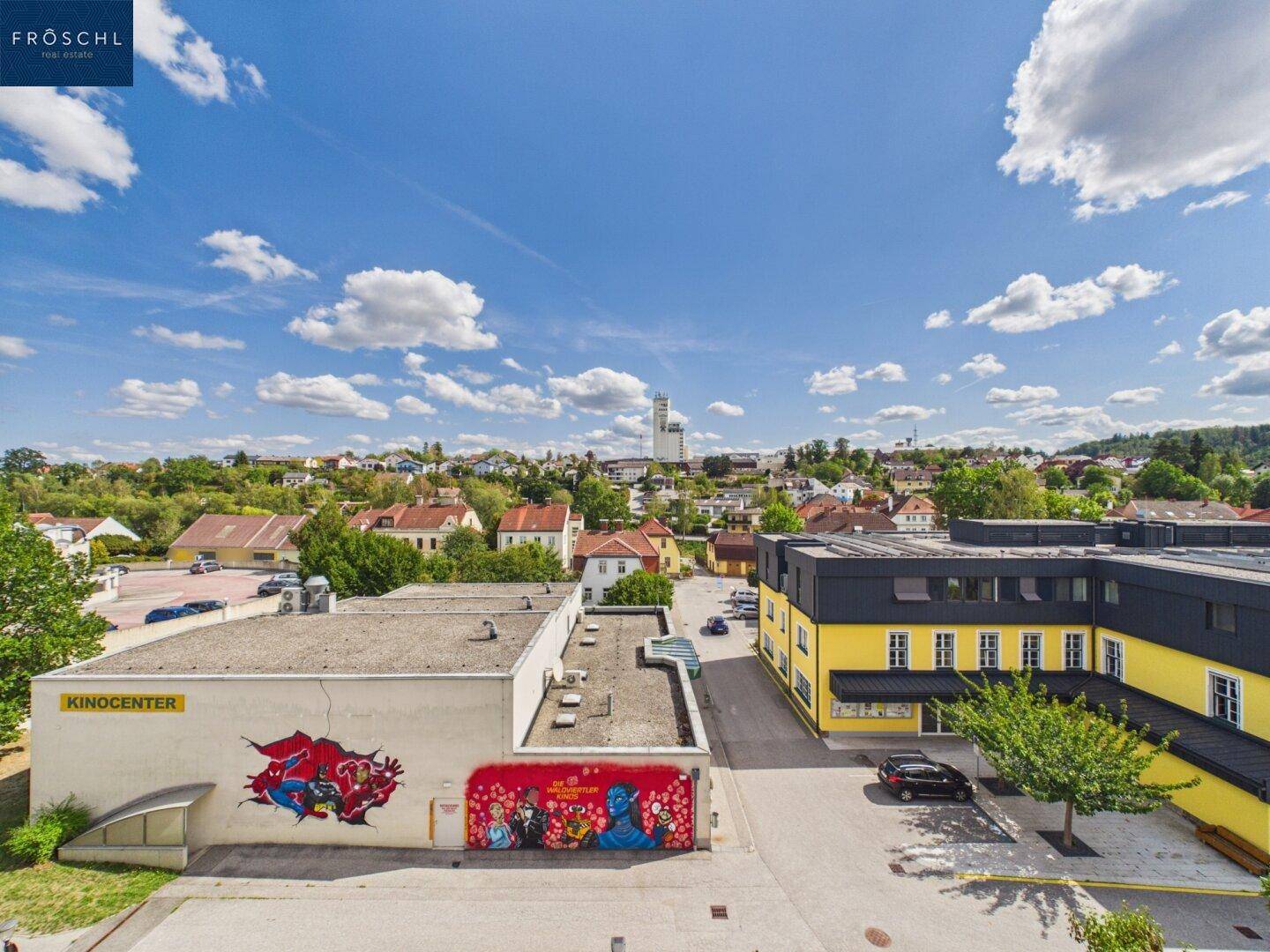 Weitläufiger Blick über eine Stadtlandschaft mit verschiedenen Gebäuden und viel Grün unter blauem Himmel.