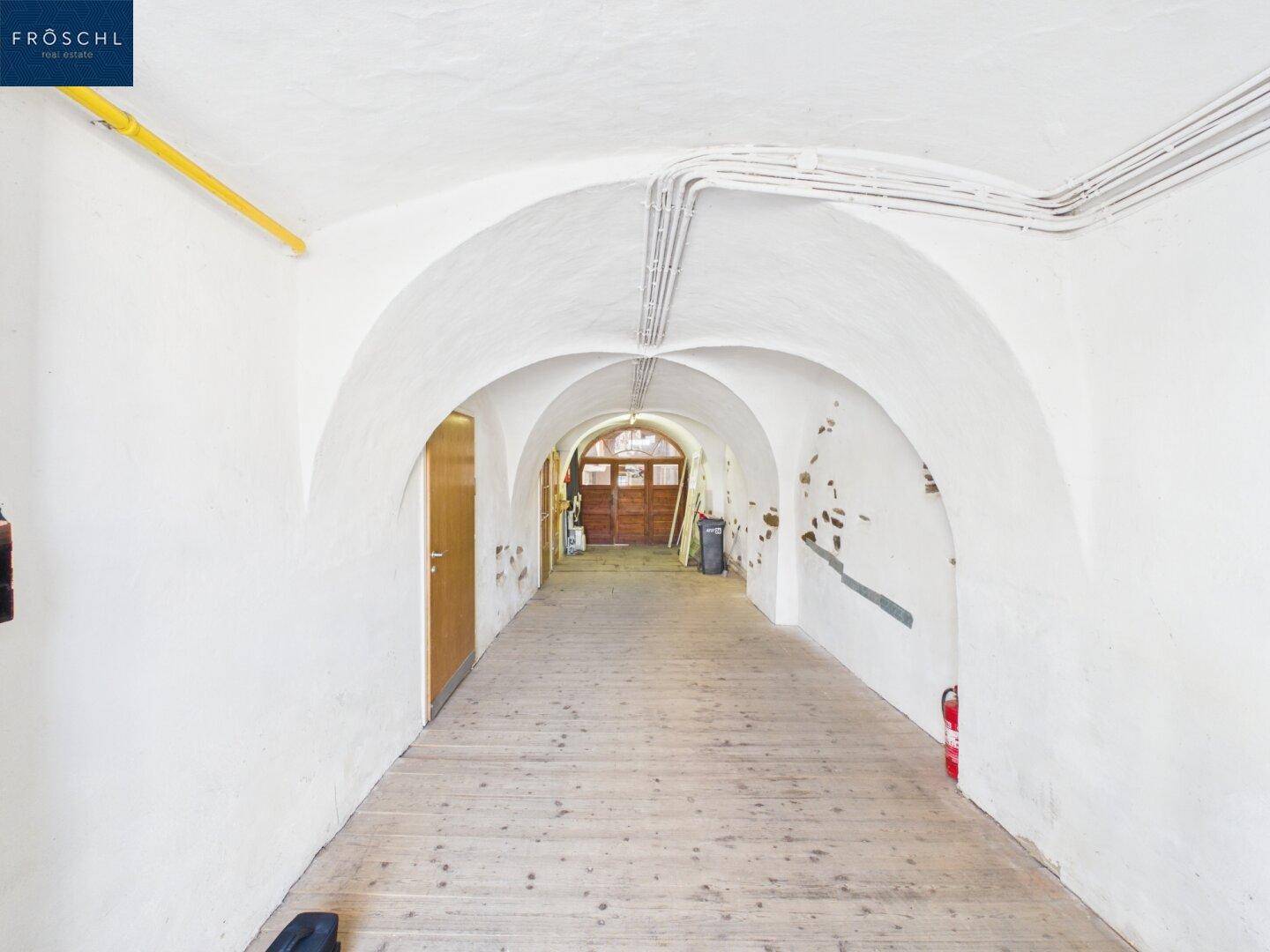 Langer, gewölbter Gang mit Holzboden und weiß getünchten Wänden, der zu einem Holztor führt.