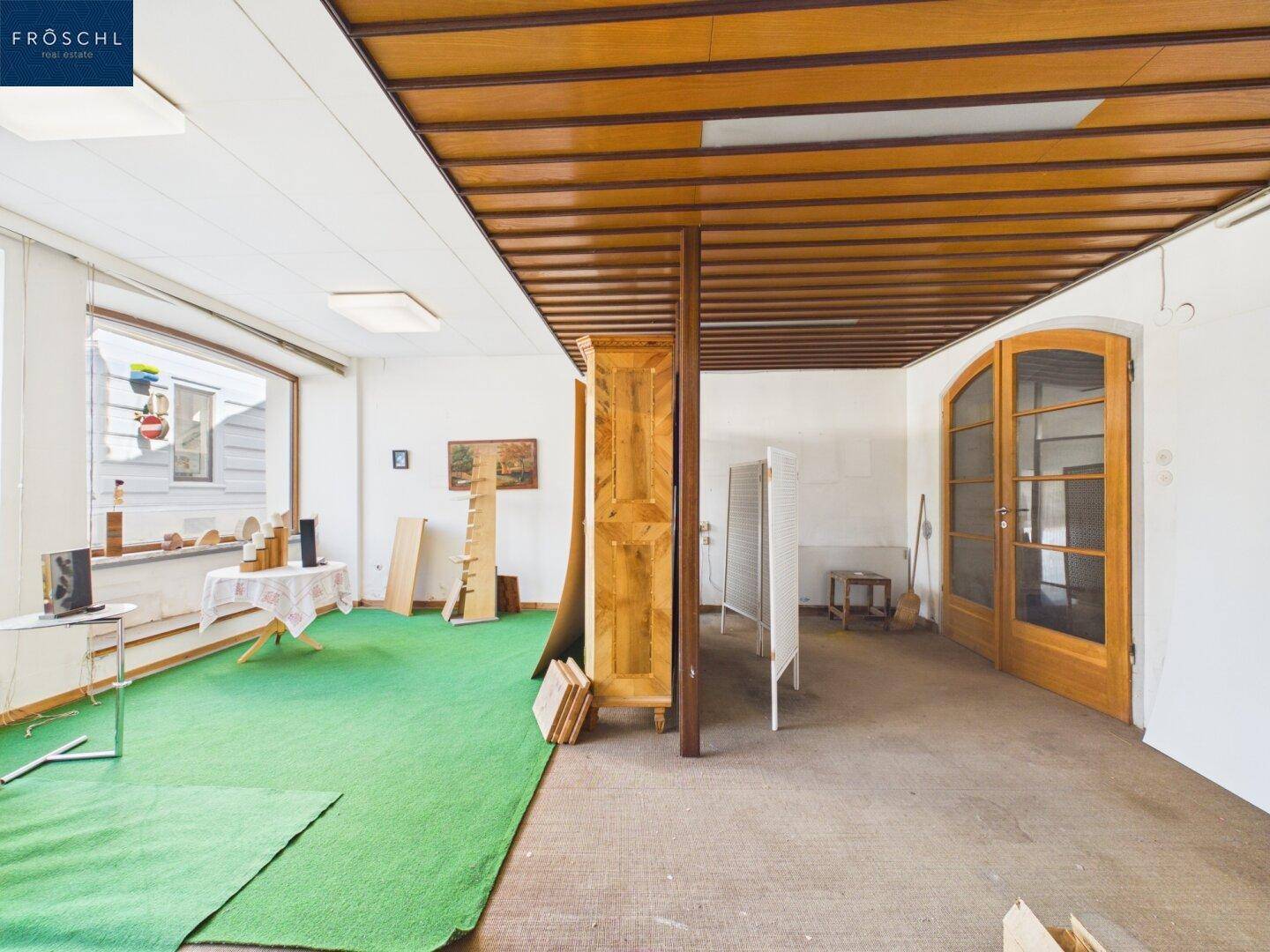 Geräumiger Innenraum mit Holzdeckenverkleidung, großen Fenstern und einer Mischung aus Teppich- und Betonboden.