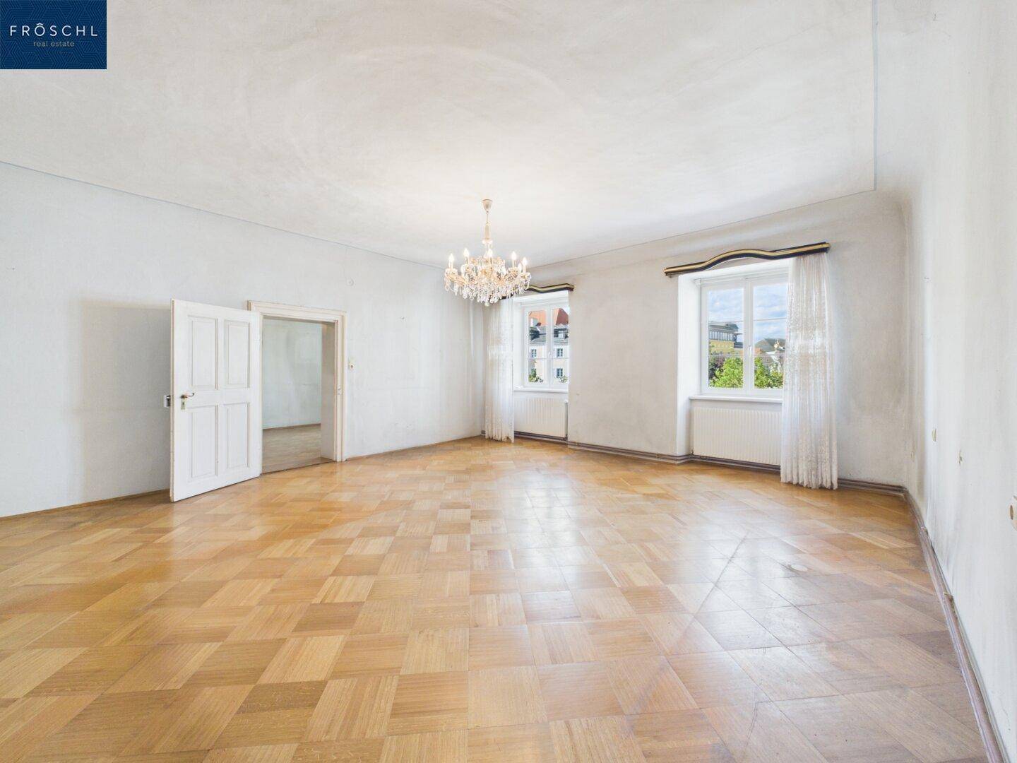 Geräumiges Wohnzimmer mit Fischgrätparkett, hohen Decken und einem eleganten Kronleuchter.