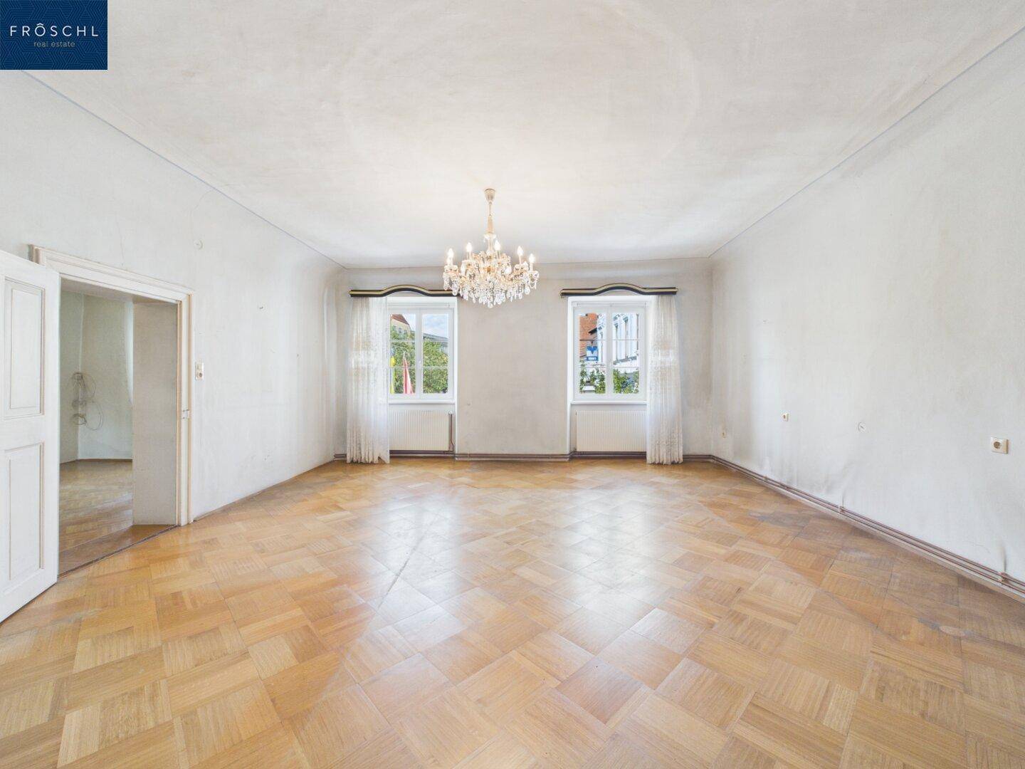 Helles Wohnzimmer mit klassischem Parkettboden, hohen Decken und einem dekorativen Kronleuchter.
