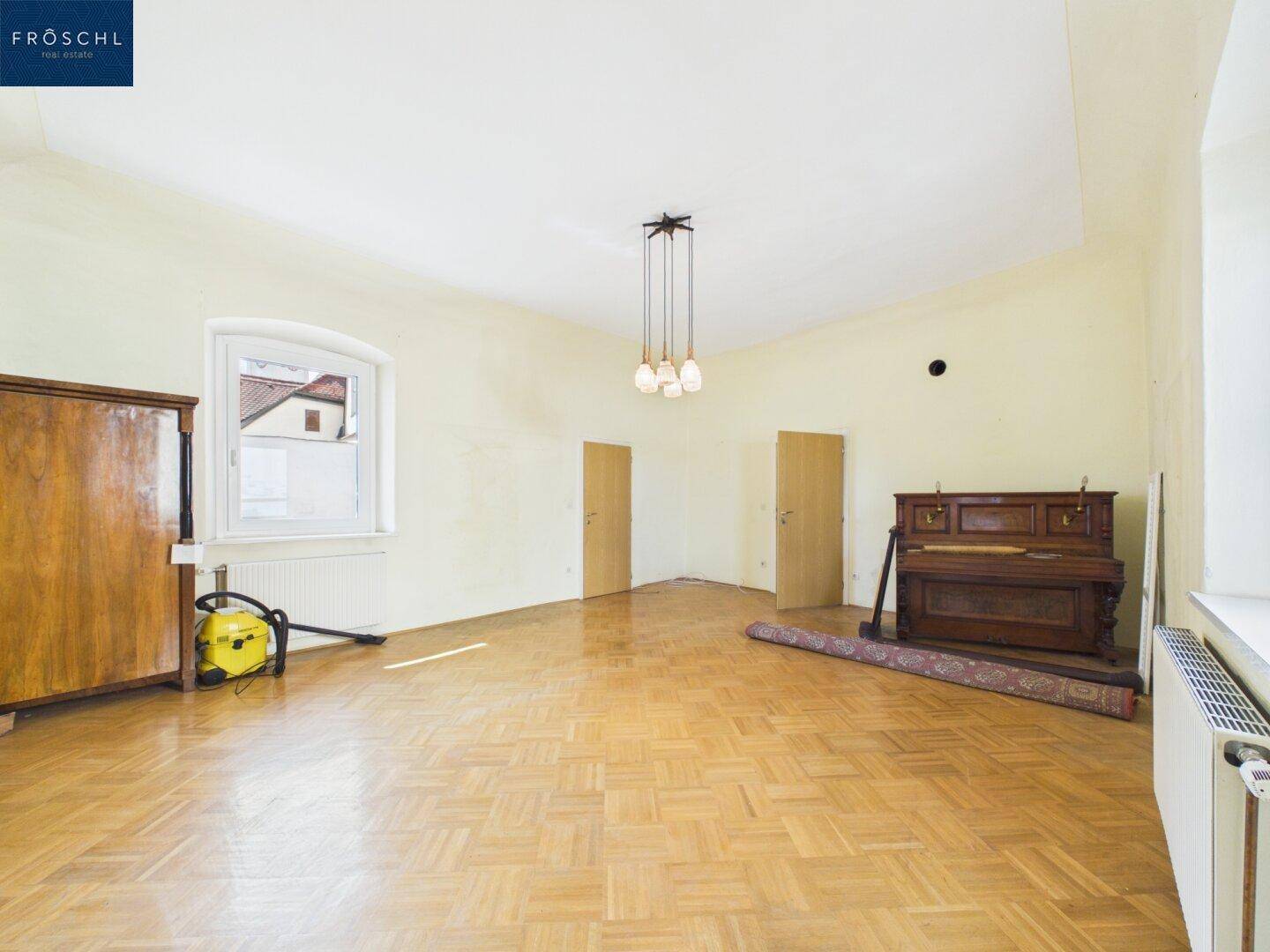 Großer Raum mit Parkettboden, hohen Decken und einem antiken Klavier und Schrank.