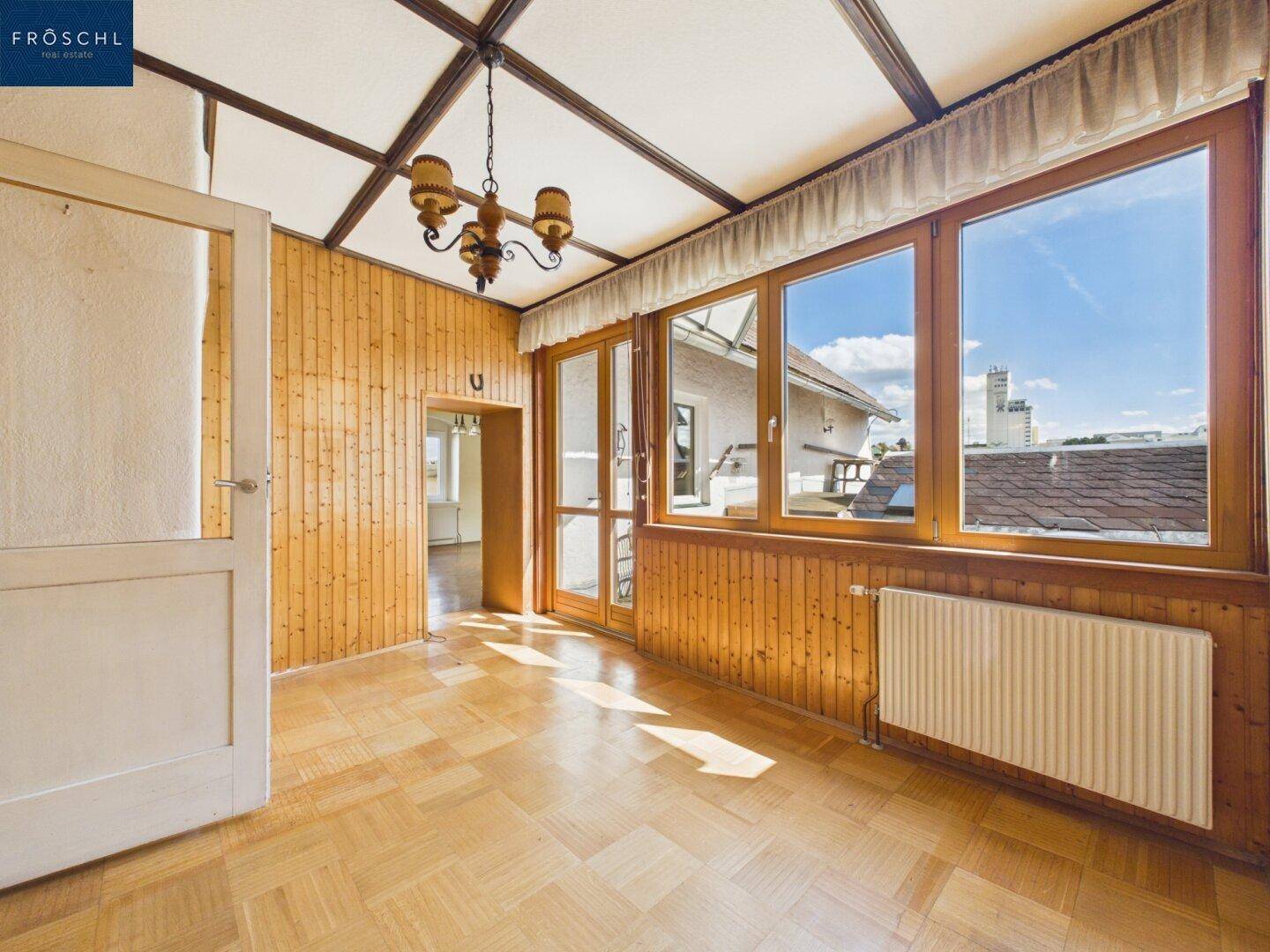 Heller Raum mit Holzvertäfelung, Parkettboden und großen Fenstern, die viel Tageslicht hereinlassen.