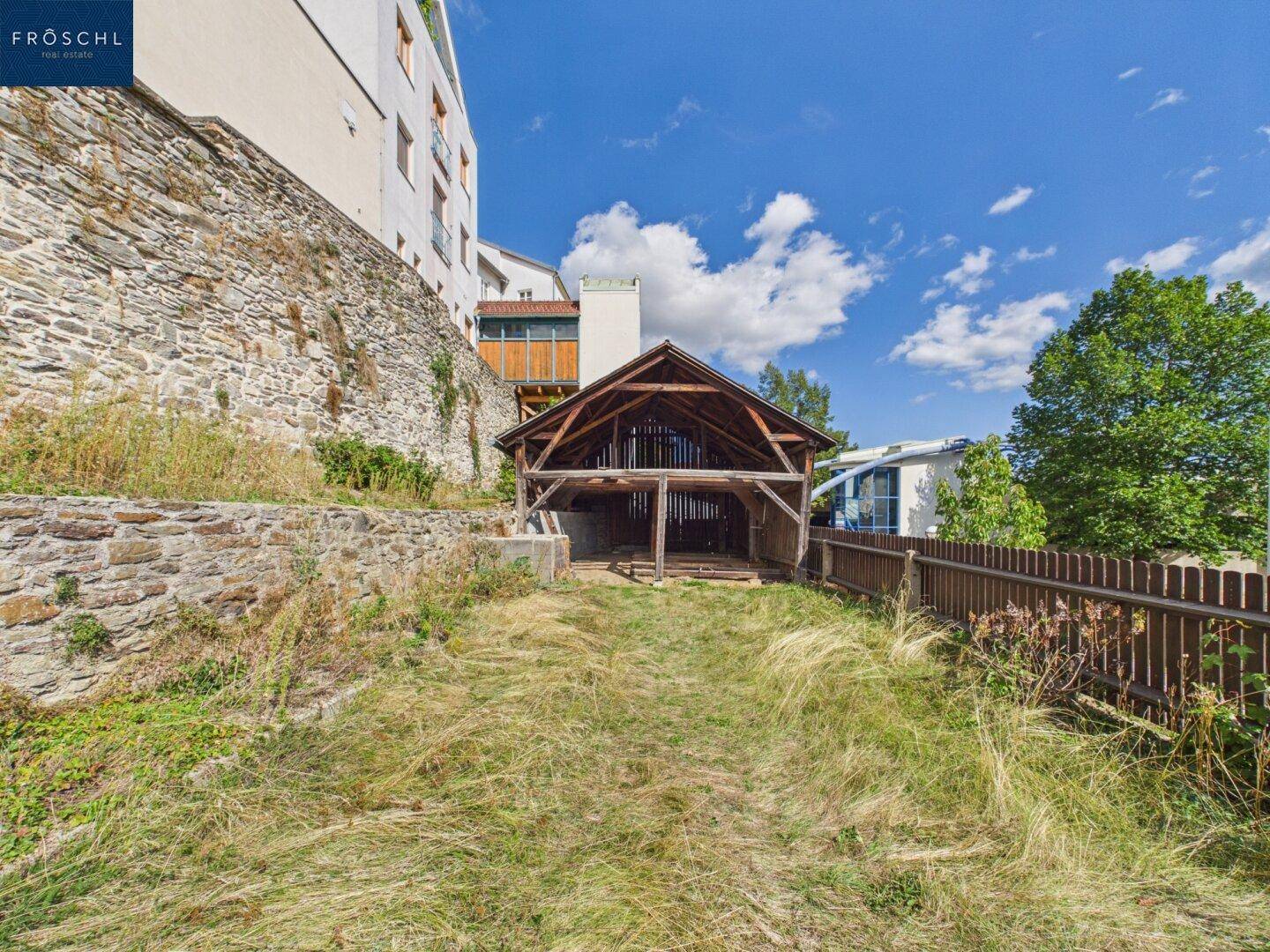 Langer, grasbewachsener Gartenbereich mit einer alten Holzscheune und einer Steinmauer im Hintergrund.